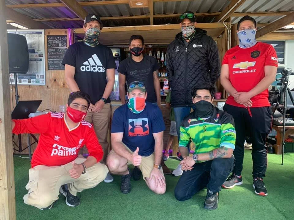 Six men wearing casual and sports clothing, all with face masks, posing inside a wooden bar or lounge area with a bar and music equipment behind them.