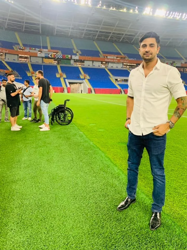 A man in a white shirt and jeans standing on a football field in a stadium. In the background, a group of people are gathered near a wheelchair.