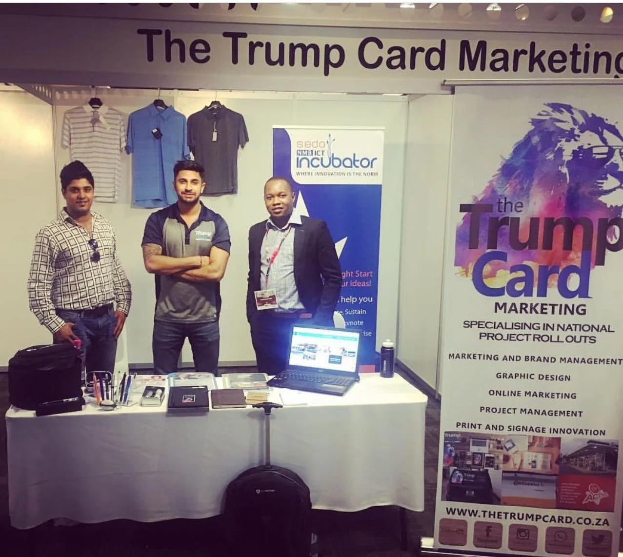 Three men standing behind a table at a marketing booth for The Trump Card, with promotional materials, notebooks, and a laptop on the table. Behind them are clothing displays and a banner with the company's services listed.