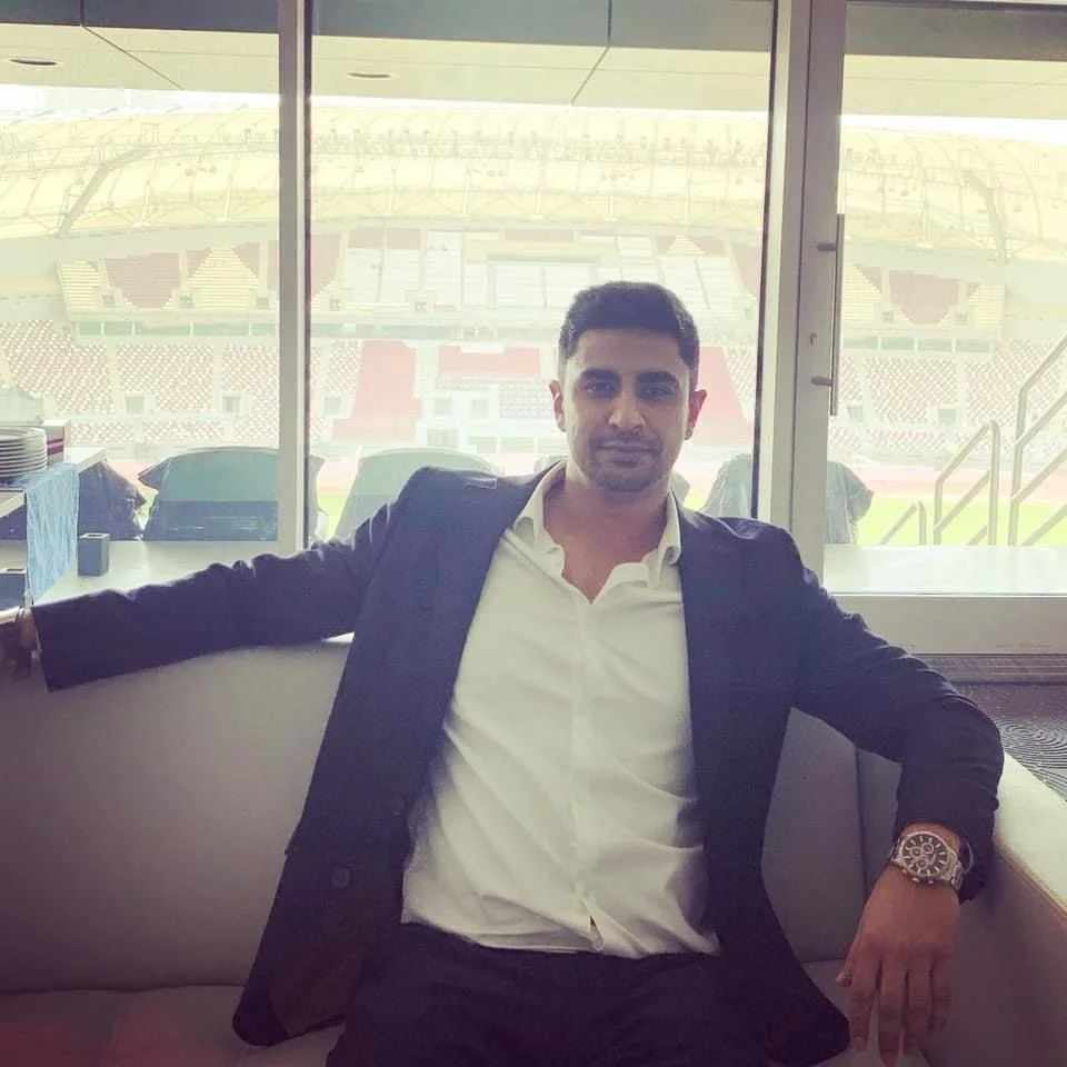 A man sitting in a stadium suite or box, wearing a suit and watch, with a stadium field visible through large window behind him.