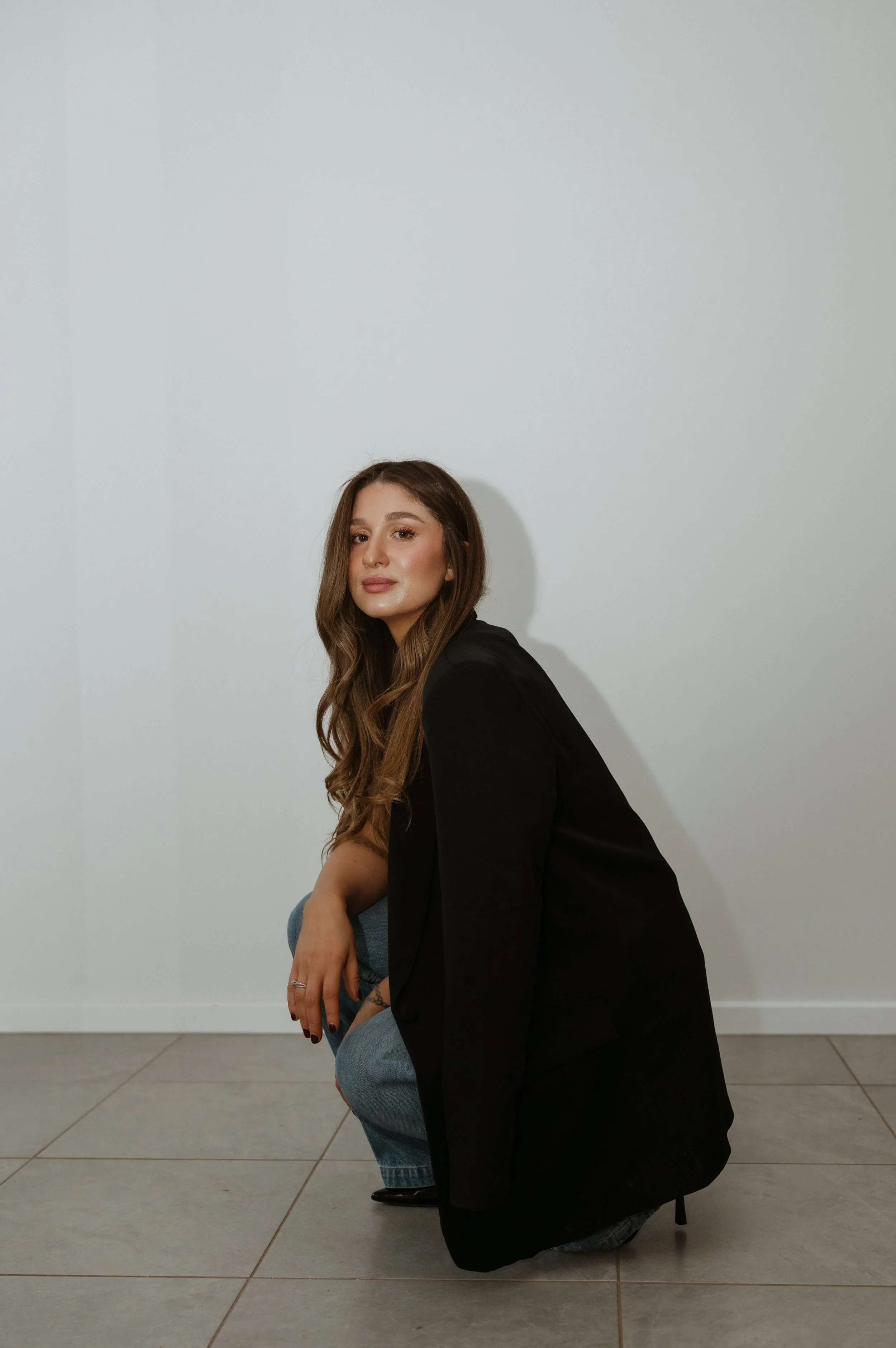 A woman with long brown hair, wearing a black blazer, is sitting on her knees on a tiled floor, with one arm resting on her knee, in front of a plain white wall. She is looking at the camera.