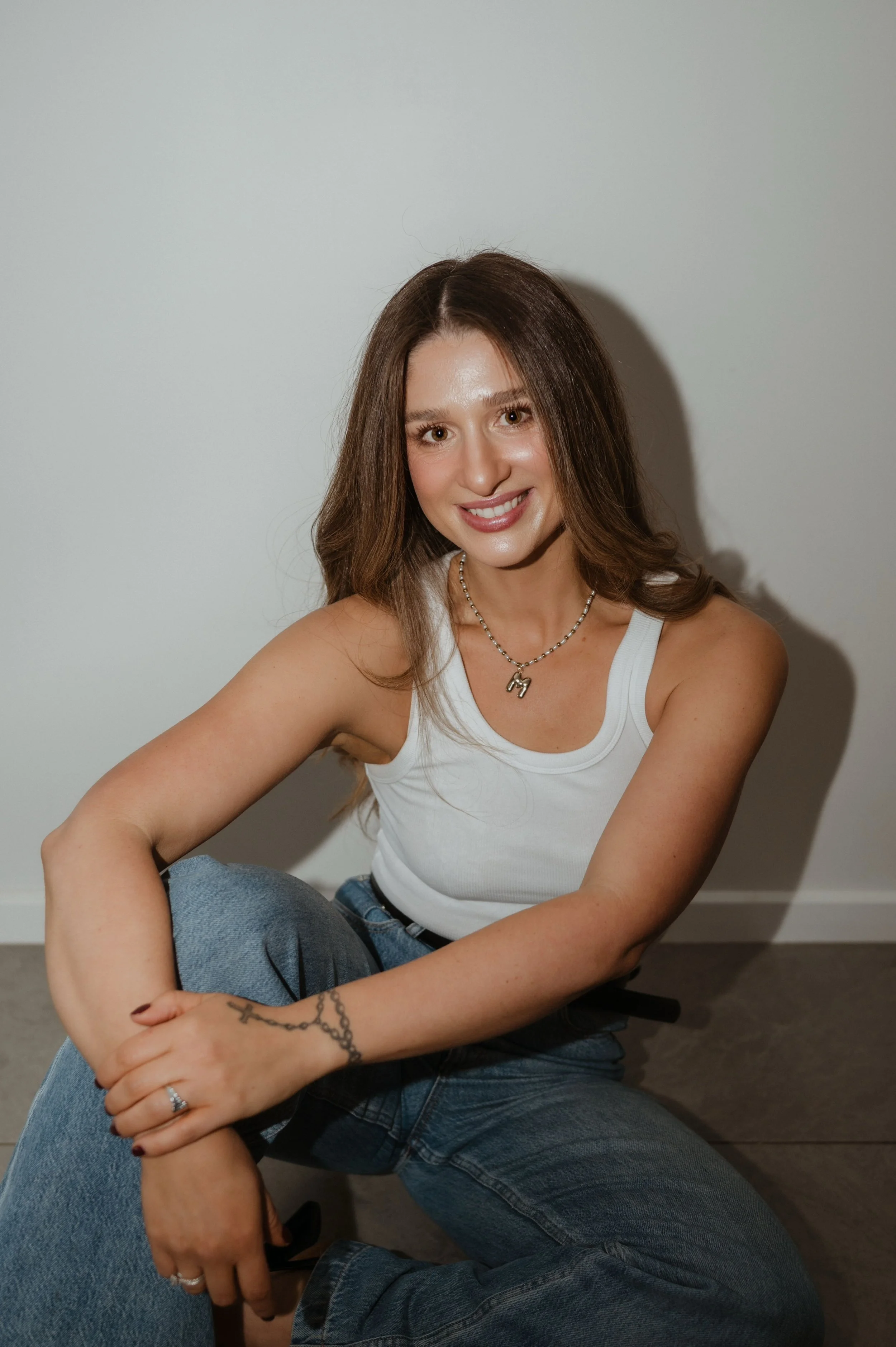 A young woman with long brown hair smiling at the camera, wearing a white tank top, blue jeans, and a necklace, sitting against a plain white wall.
