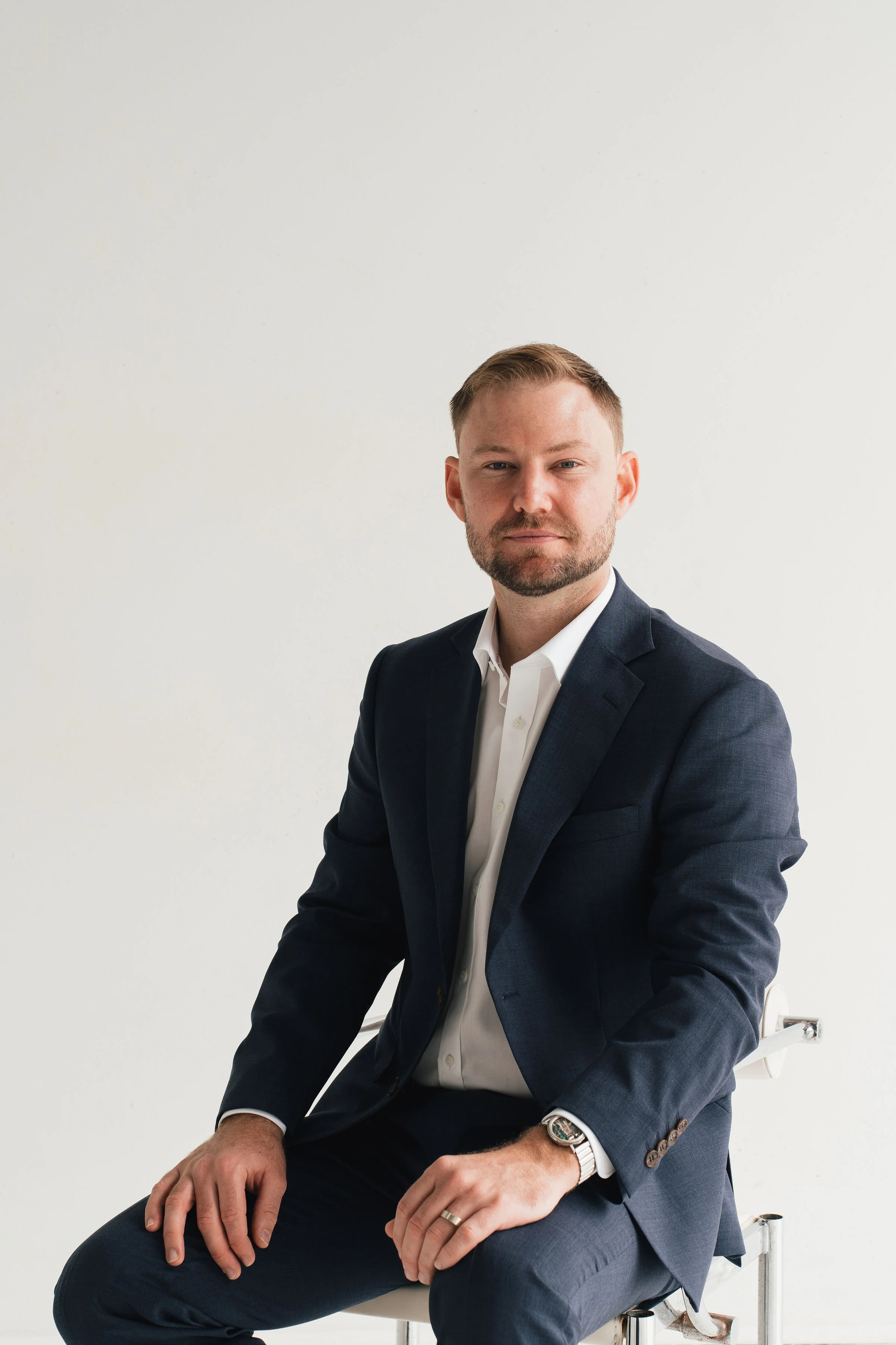 A man in a navy suit and white dress shirt sitting on a white chair against a plain white background.