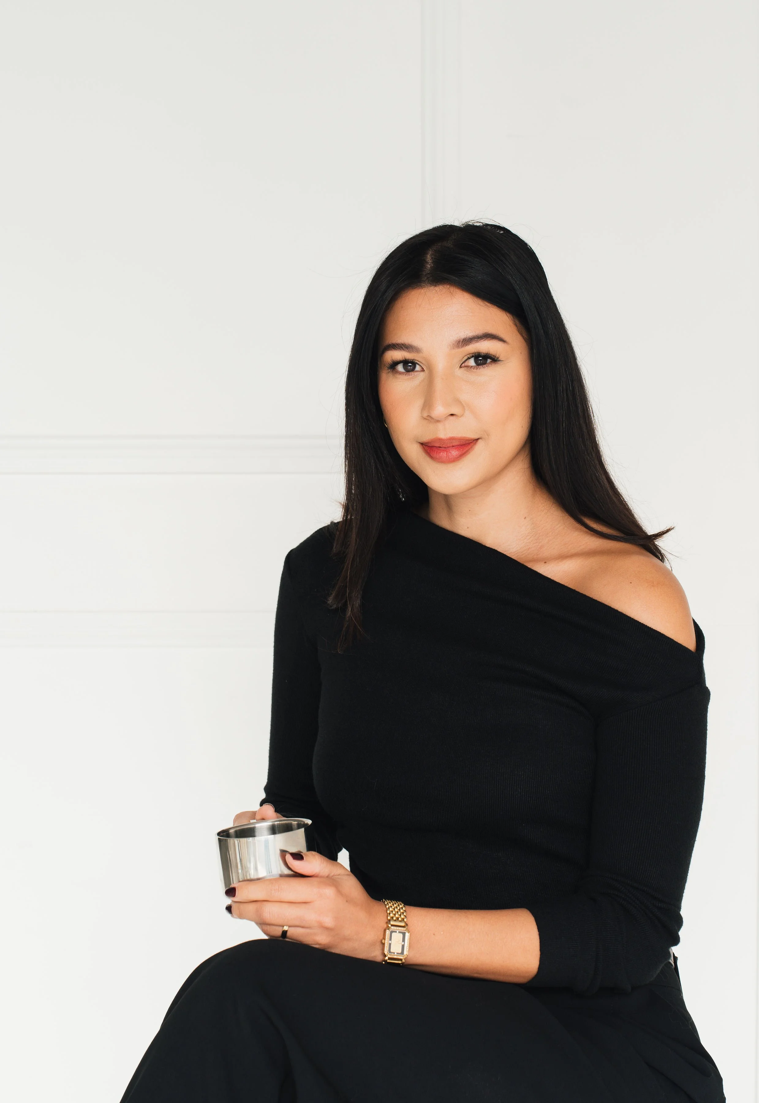A woman with long black hair, wearing a black off-the-shoulder top and black pants, holding a stainless steel cup, sitting against a white background.