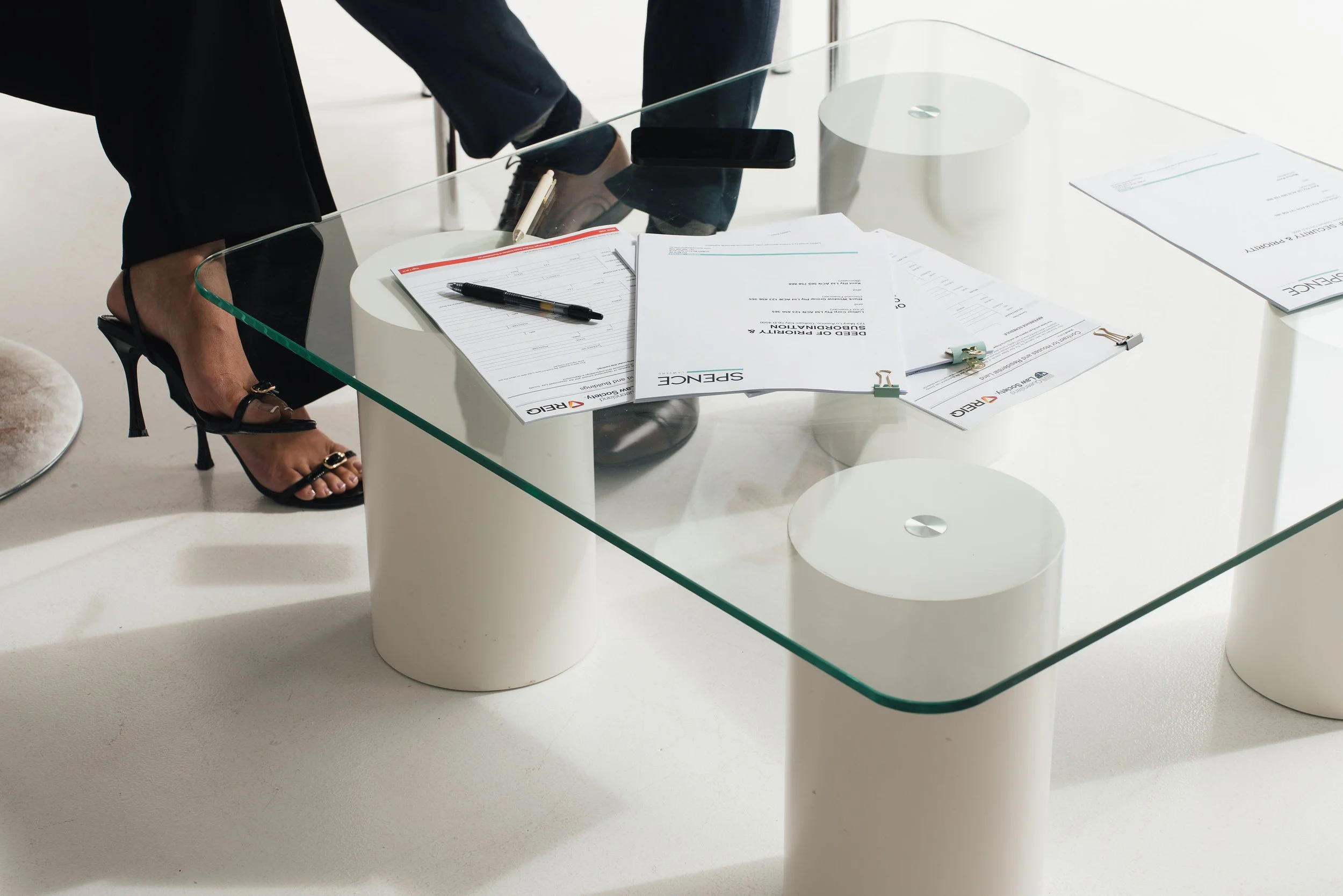 A glass conference table with documents, a pen, and a smartphone on it, with someone in black high heels standing nearby.