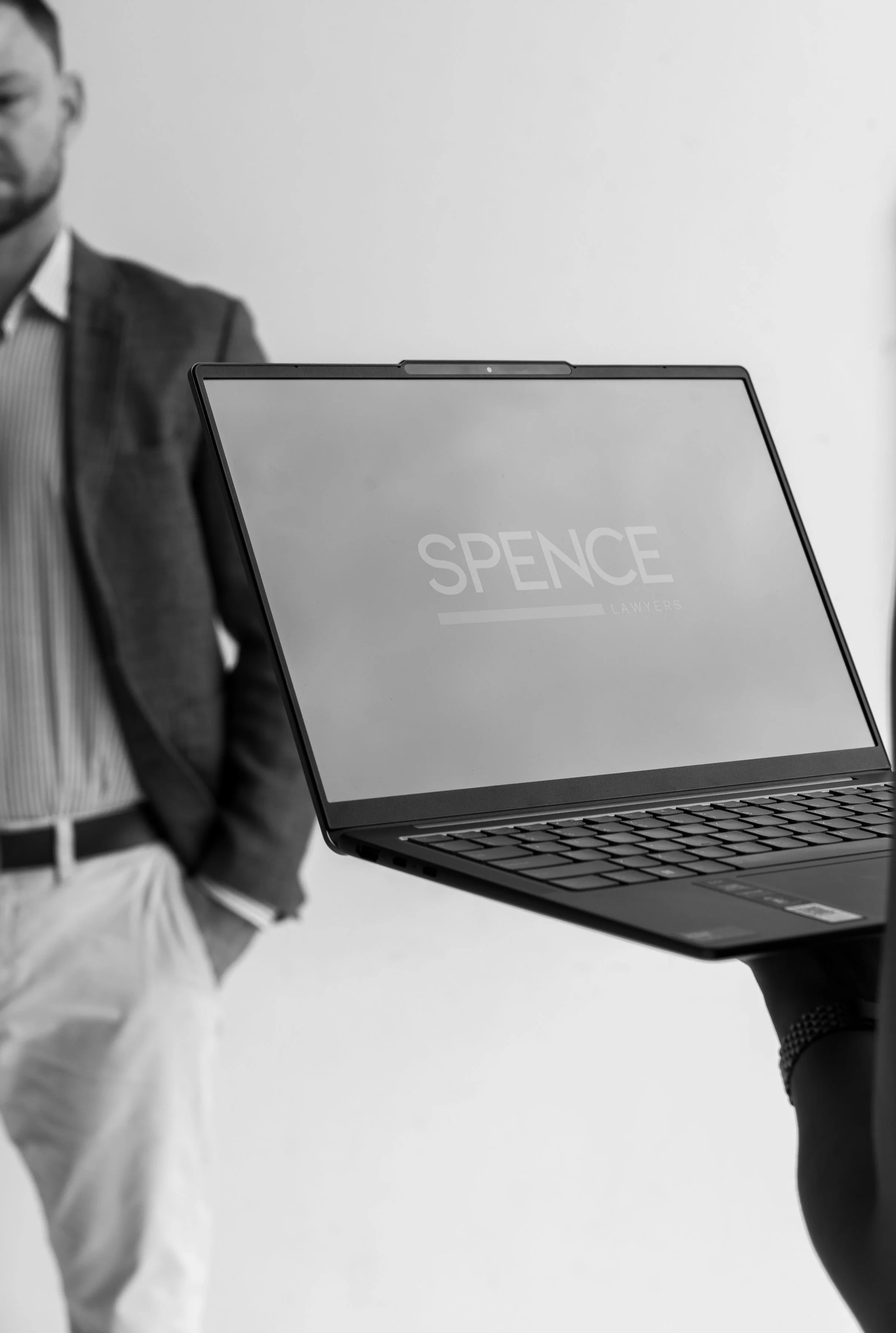 A person standing with their hand in their pocket and a laptop with the logo 'Spence Lawyers' on the screen in front of them, in black and white.