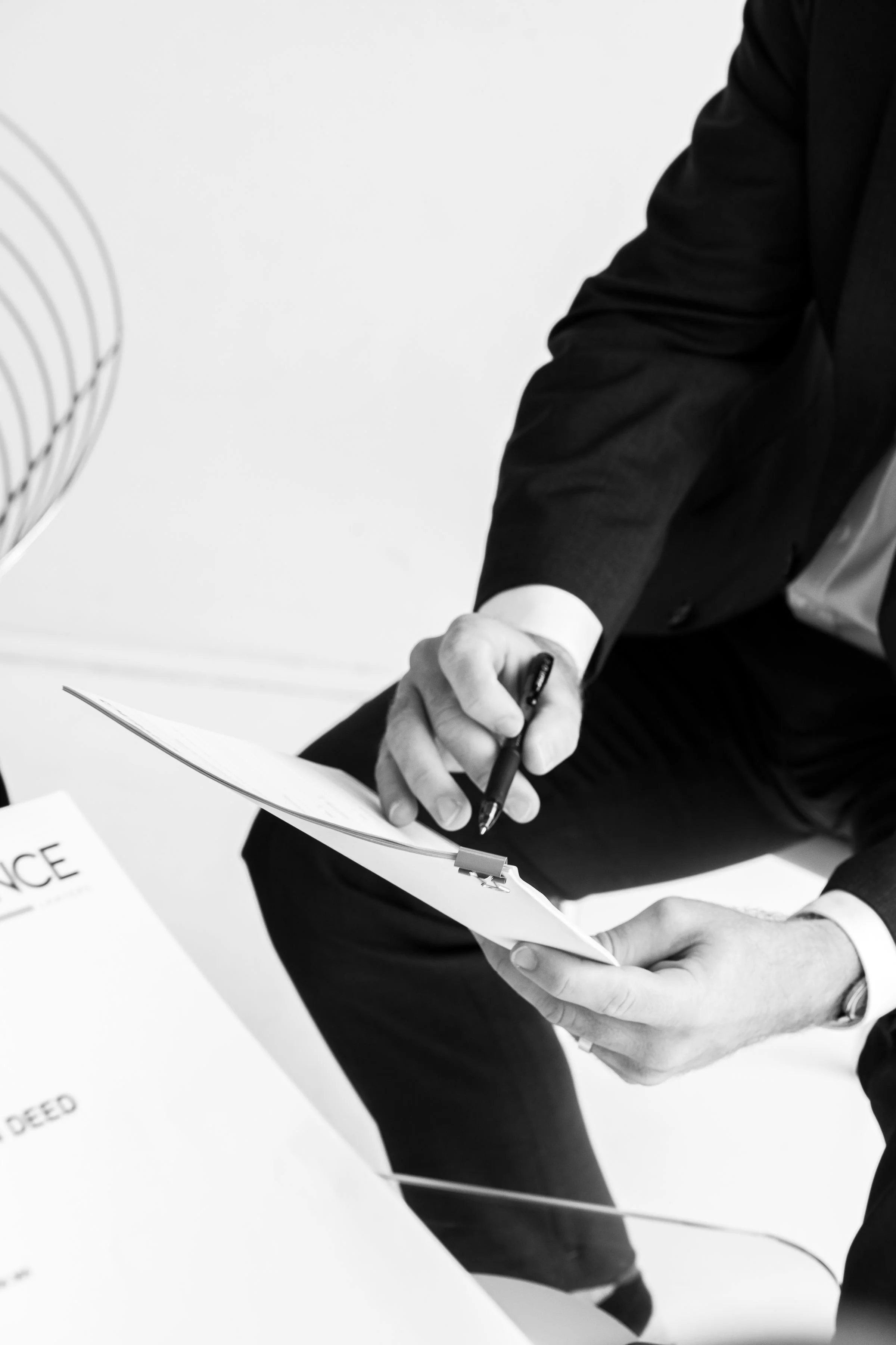 A person in a black suit sitting and holding a clipboard with a pen, appearing to review or sign a document, in an office setting.