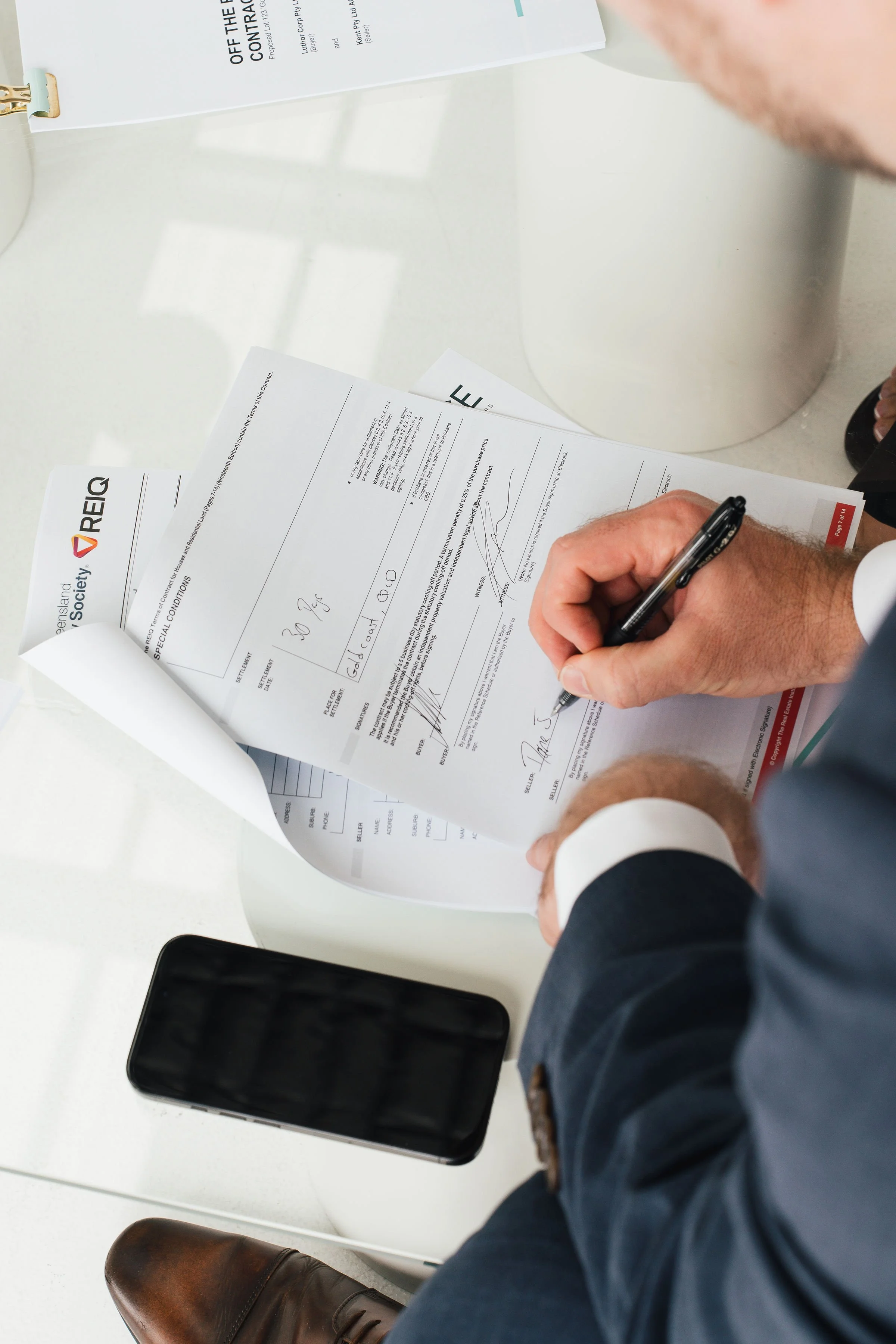 A man in a business suit signing a document at a desk with a smartphone nearby, papers and a white box are also on the desk.