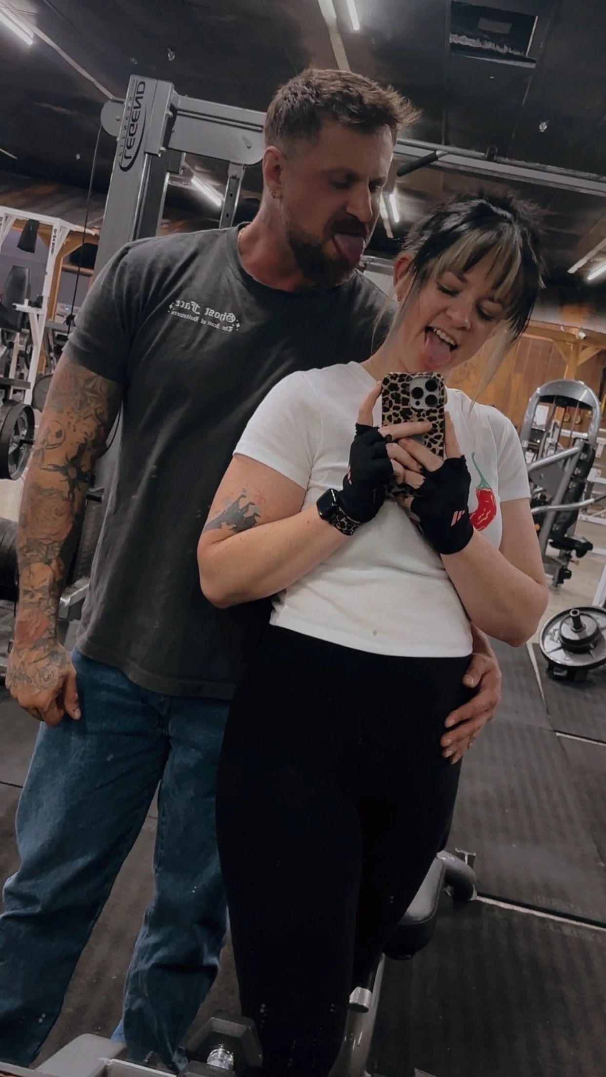 A couple taking a mirror selfie at the gym, both sticking out their tongues. The woman is holding the phone with a leopard print case, and the man is hugging her from behind, with his hand on her waist.
