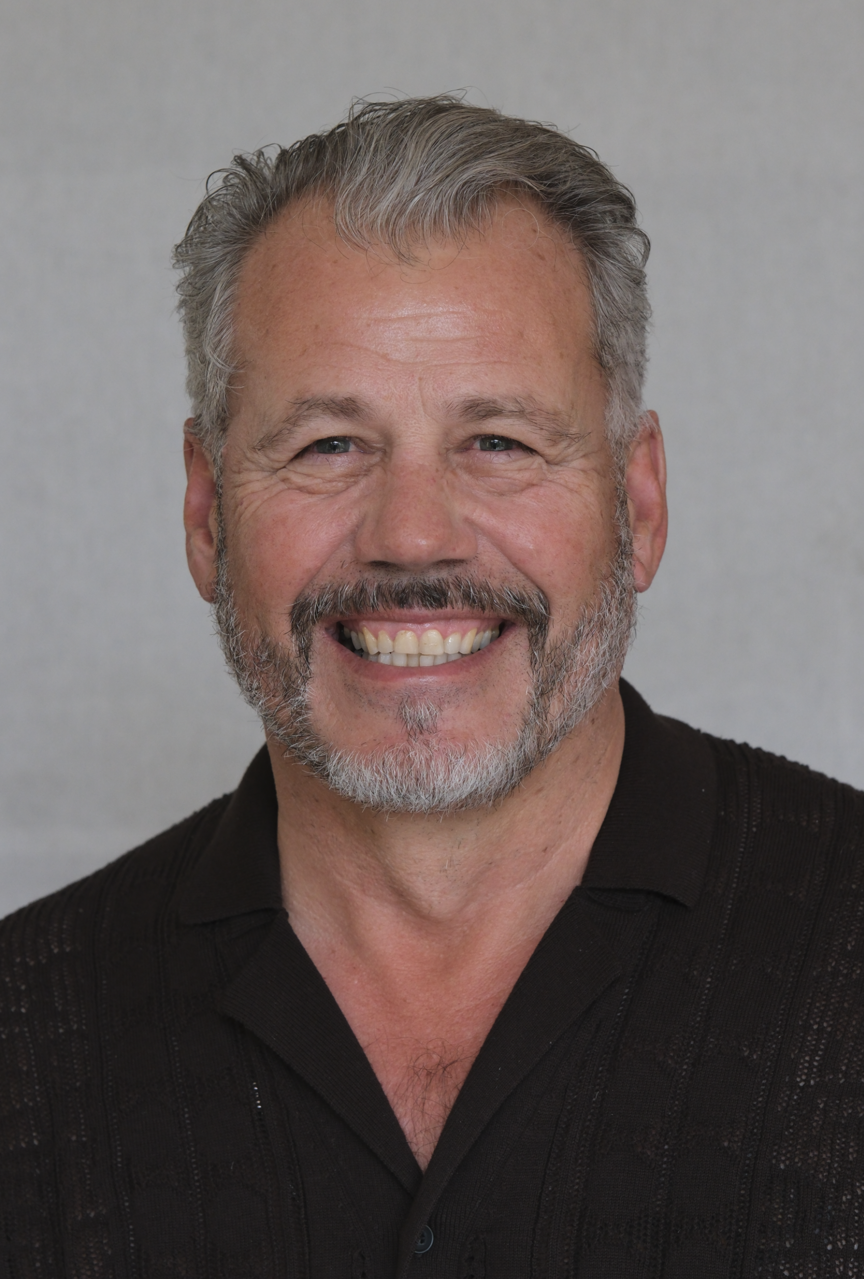 A middle-aged man with gray hair and beard smiling, wearing a black button-up shirt, in front of a plain gray background.
