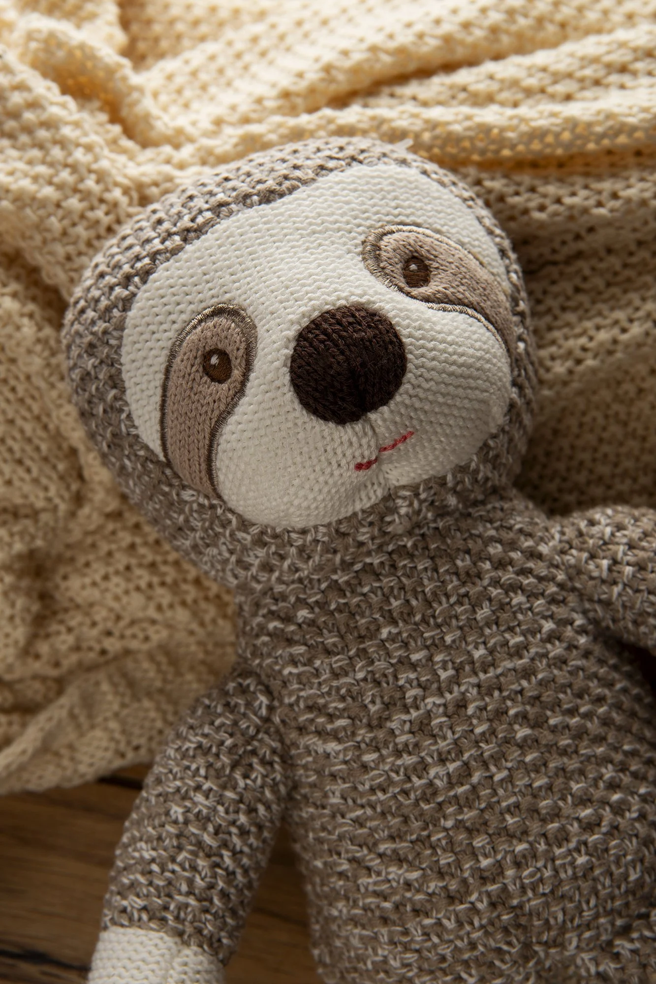 A knitted stuffed animal resembling a sloth, with a detailed face, lying on a soft, beige fabric background.