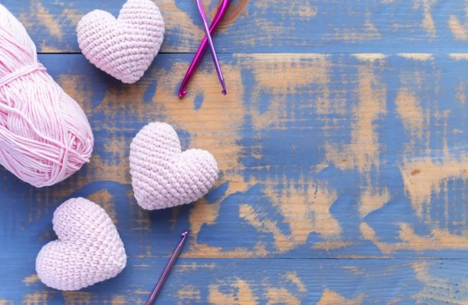 Pink crochet hearts, embroidery needles, pink yarn, and a pink crocheted heart on a distressed blue wooden surface.