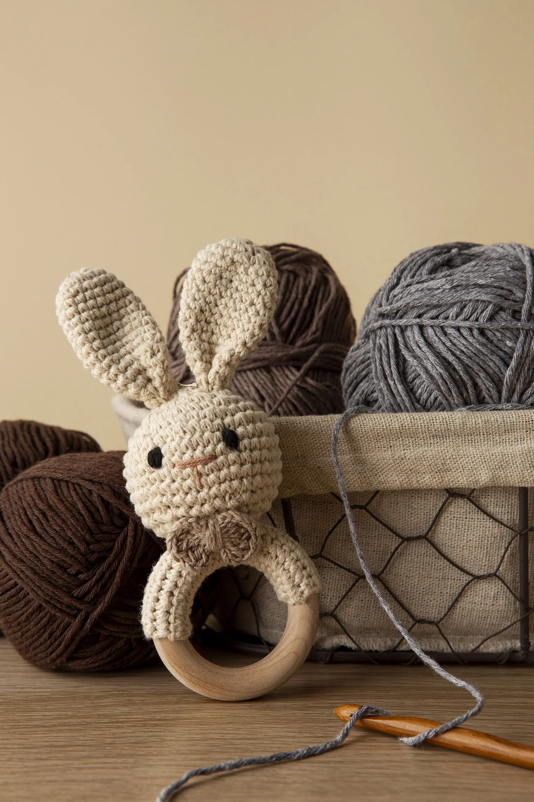 A crocheted bunny toy with big ears and a wooden ring body, surrounded by balls of brown and gray yarn in a fabric basket, on a wooden surface against a plain beige wall.