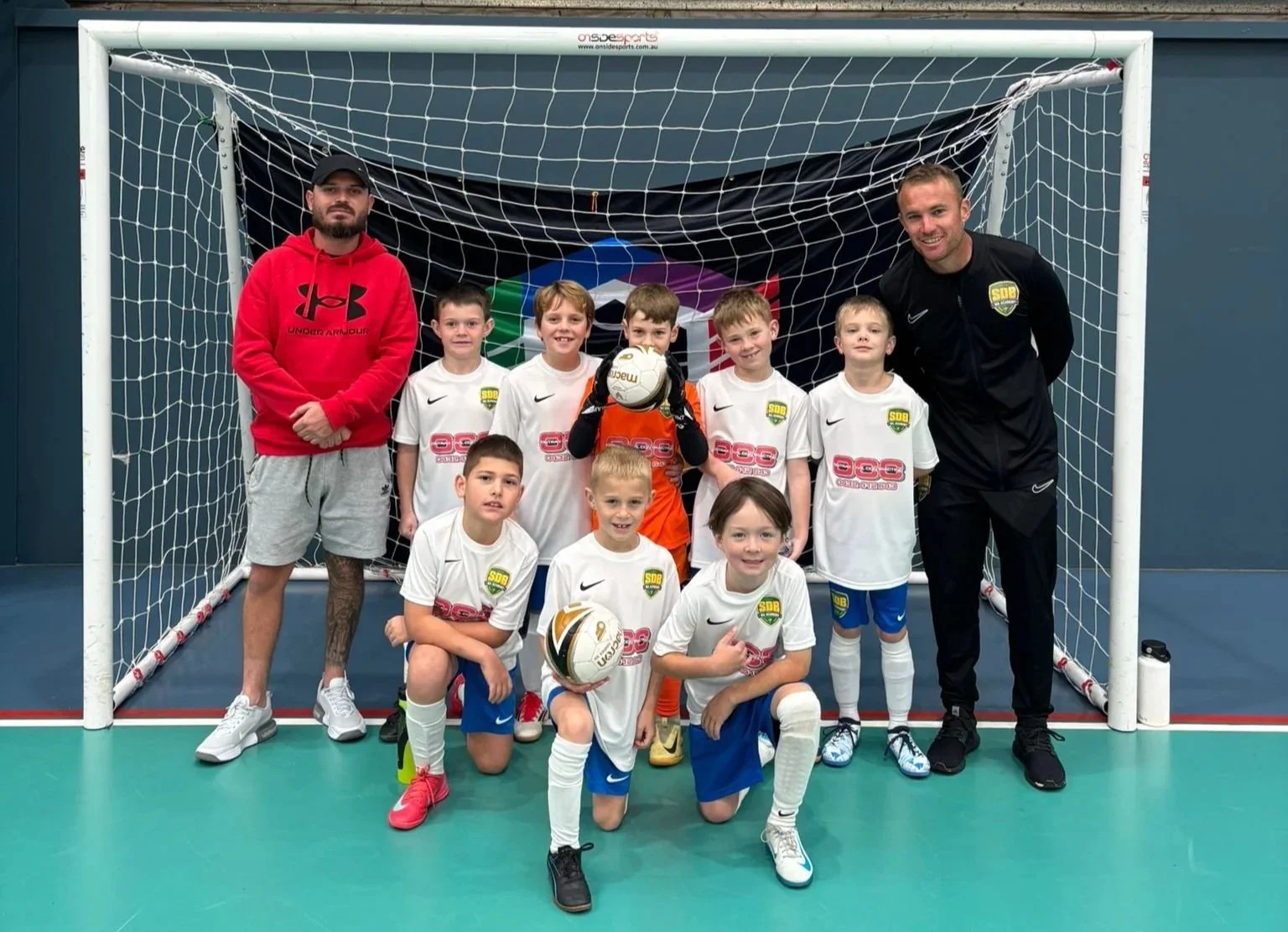 A youth indoor soccer team with 9 young boys and 2 coaches posing inside a goal on a green court. The boys are in white jerseys and blue shorts, some holding soccer balls, and there is a multicolored logo on the goal backdrop.