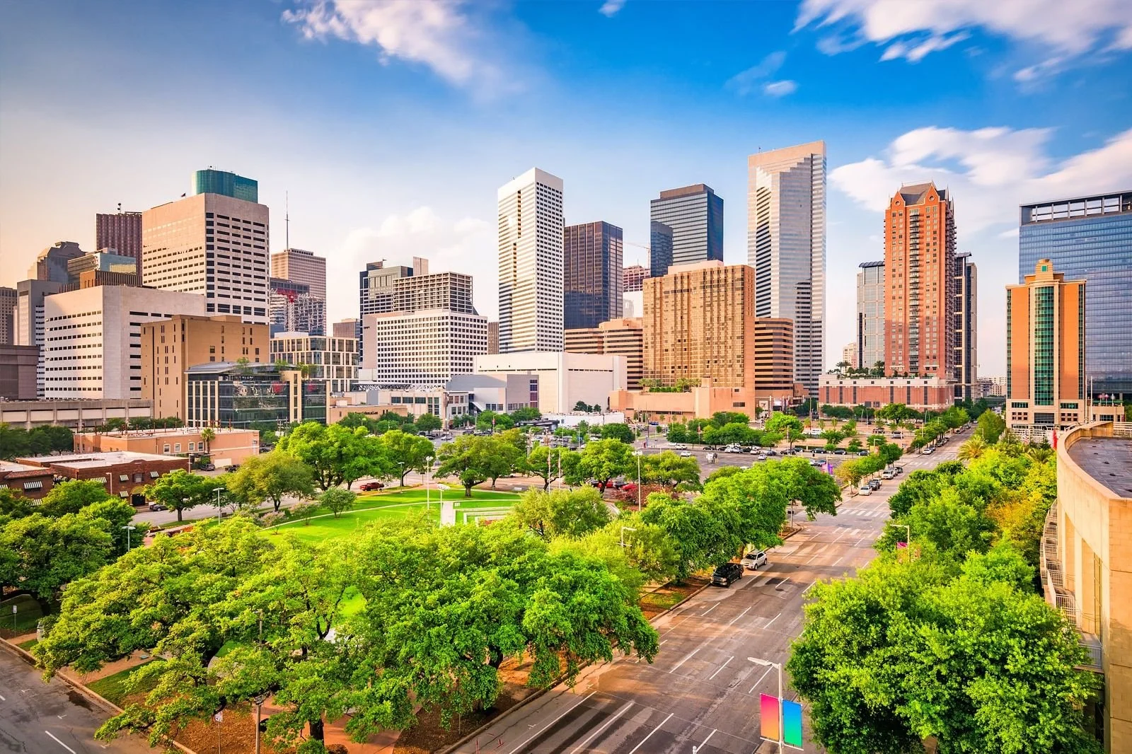 Downtown cityscape with tall skyscrapers, green trees, and a parking lot under a partly cloudy sky.