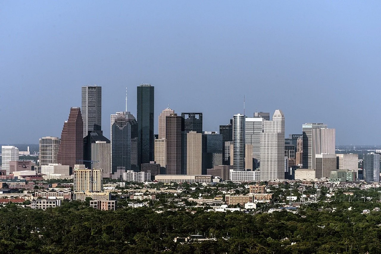 City skyline with tall skyscrapers against a clear blue sky and green trees in the foreground.