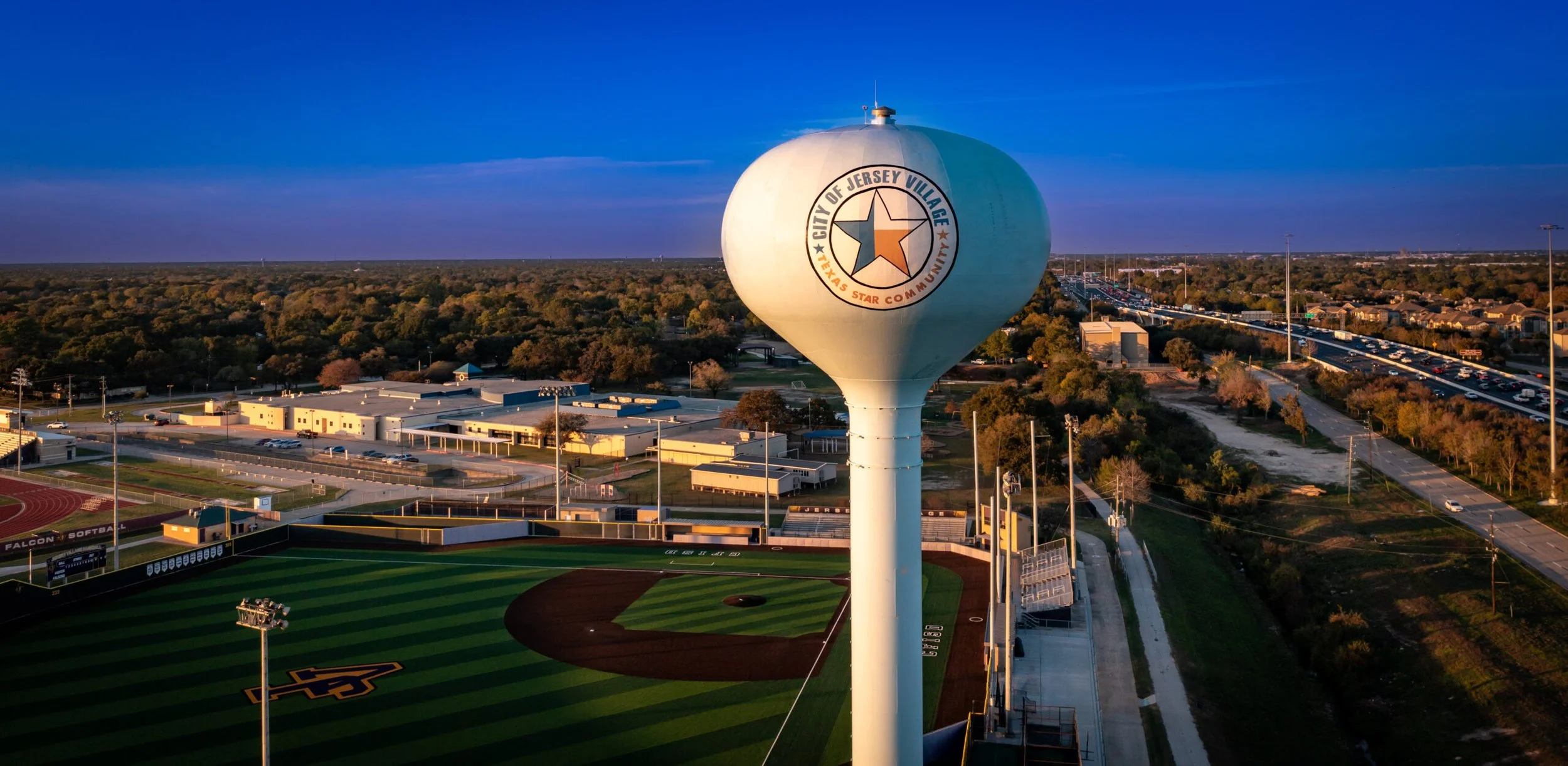 A water tower with the logo of Jersey Village, Texas, above a baseball field at a sports complex at sunset.