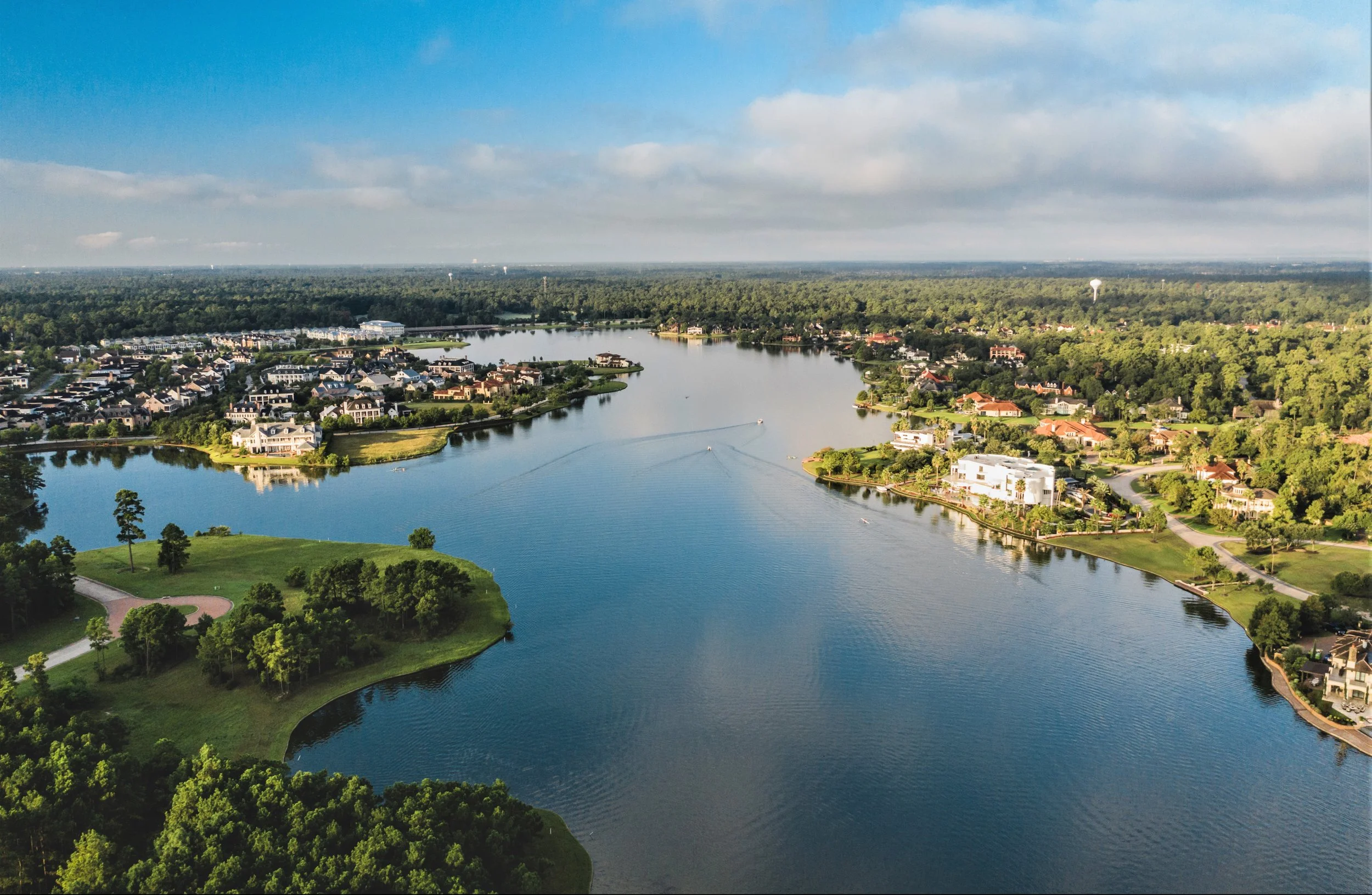 Aerial view of a large lake surrounded by residential neighborhoods and greenery, with houses, boats on the water, and trees along the shoreline.