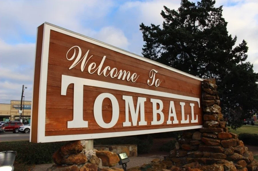 Sign that says "Welcome To TOMBALL," mounted on stone supports with a background of trees and a parking lot.