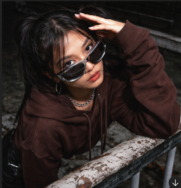 A young woman with black hair, wearing dark sunglasses, a brown hoodie, and silver jewelry, resting her elbow on a rusted metal railing outdoors, looking at the camera.