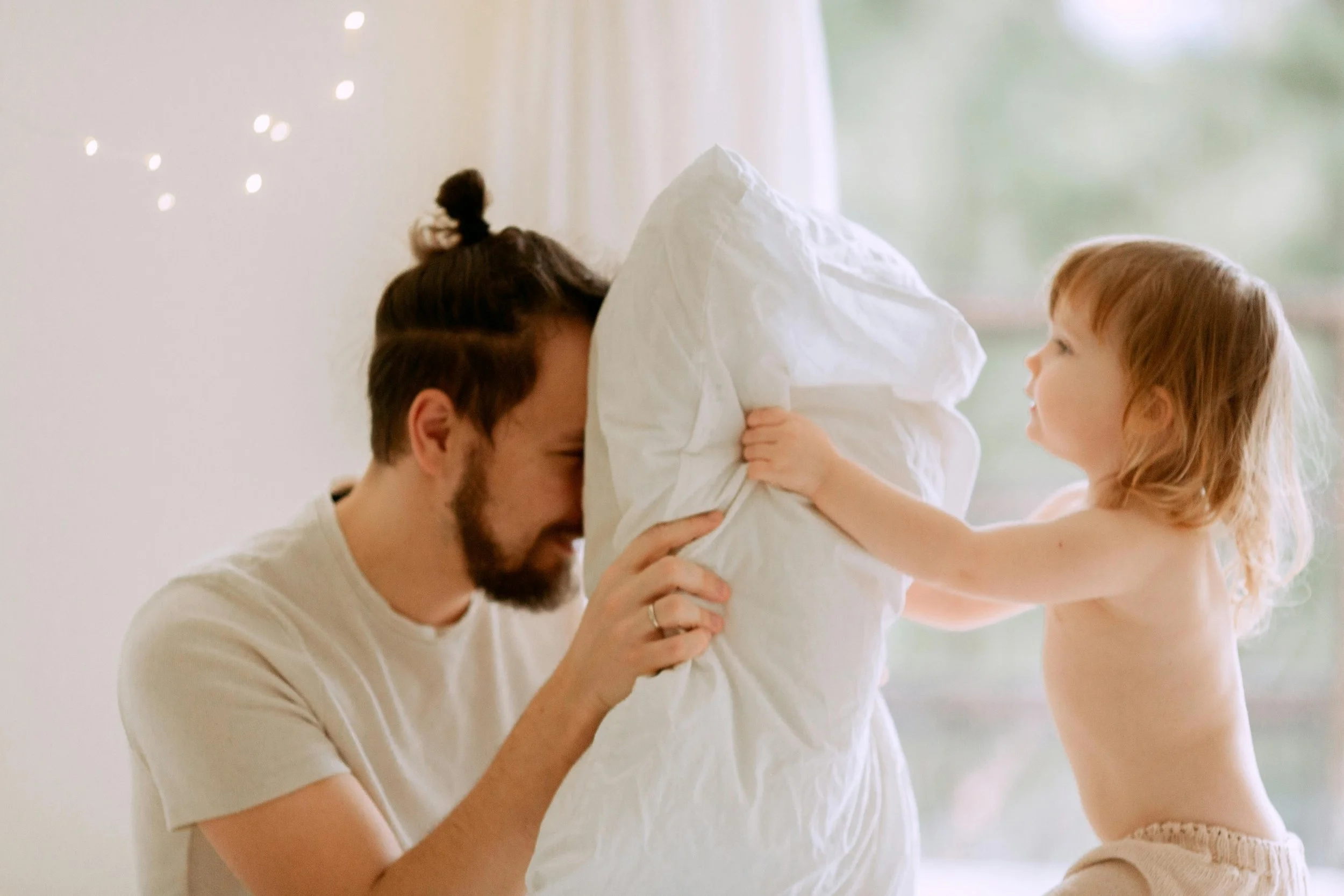 A father and daughter playfully holding a pillow together in a bright room, smiling and enjoying a moment of bonding.