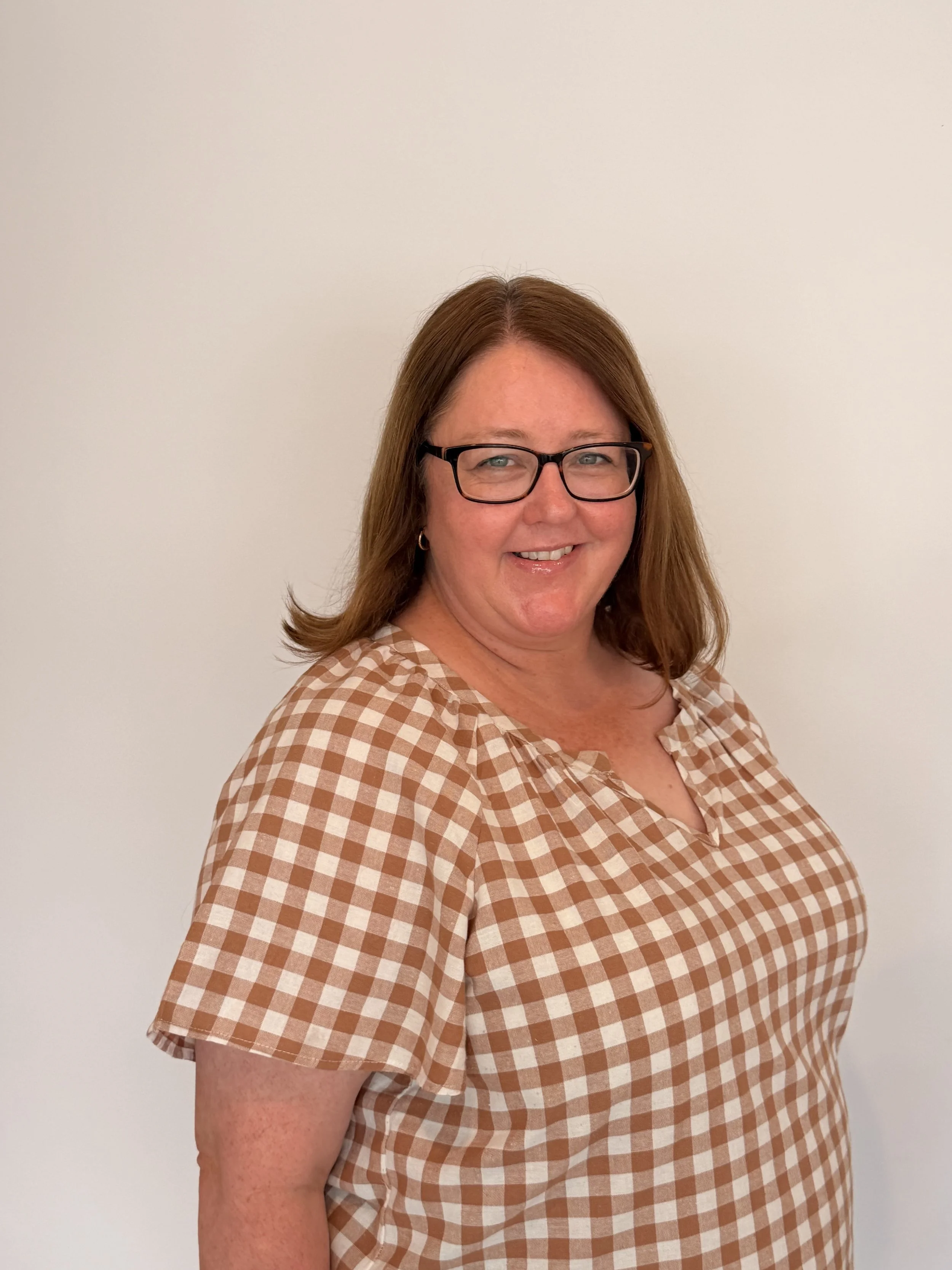 A woman with red hair, glasses, and earrings wearing a brown and white checkered shirt, smiling while standing against a plain white wall.
