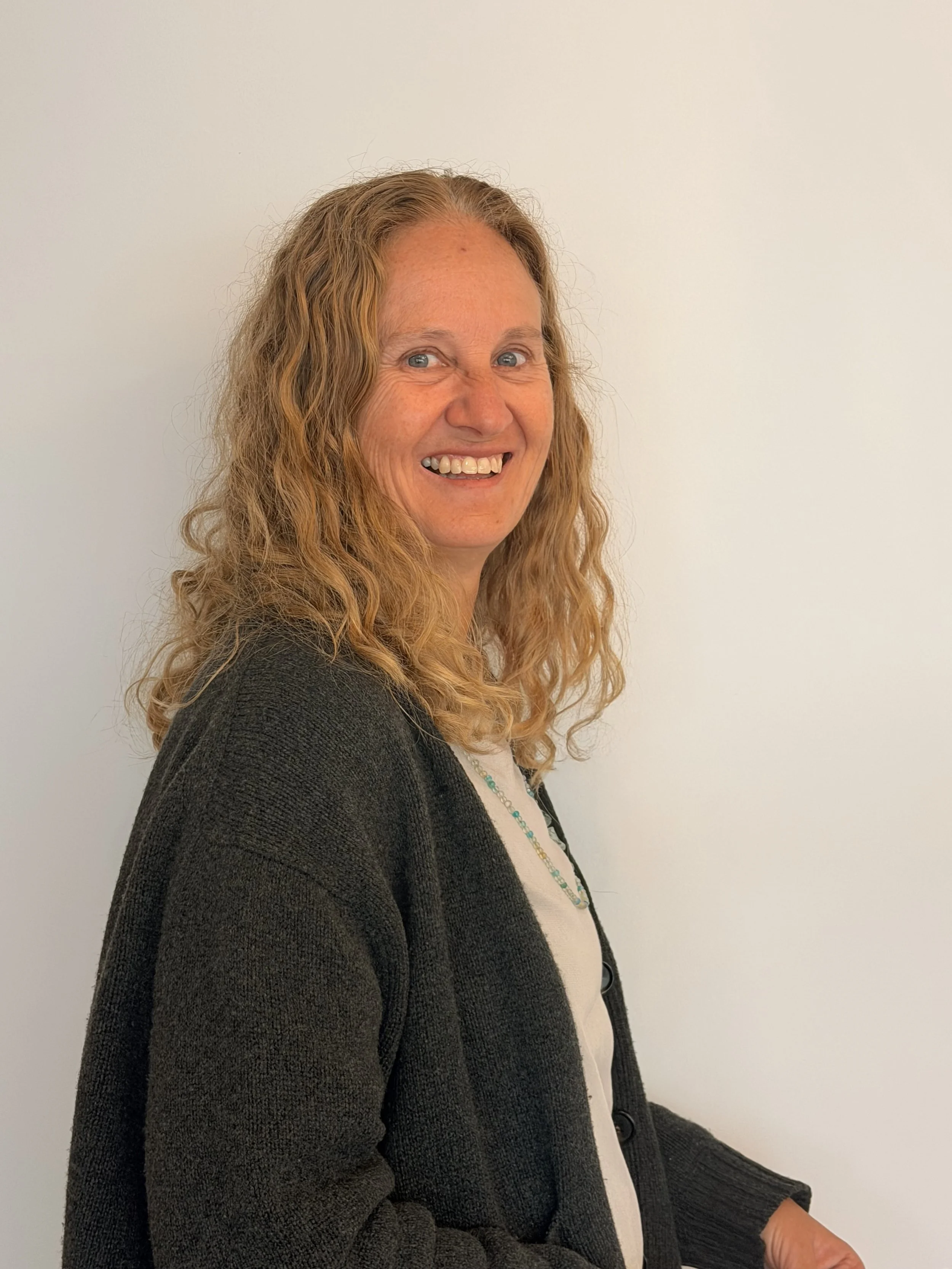 A woman with curly red hair smiling and looking at the camera, wearing a dark sweater and a beaded necklace, standing against a plain white wall.