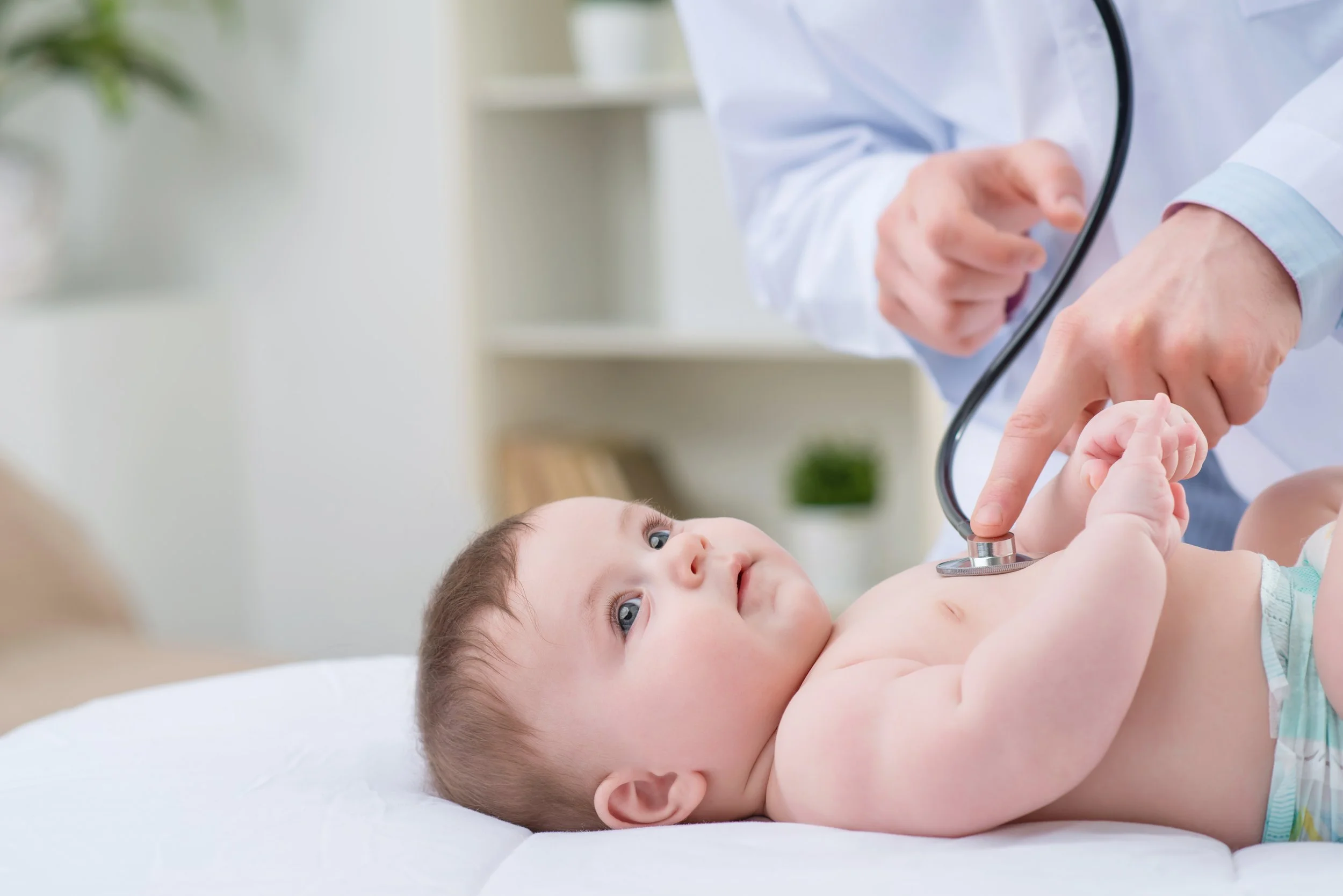 A young child lying on a hospital bed being examined by a doctor with a stethoscope, in a bright medical clinic.