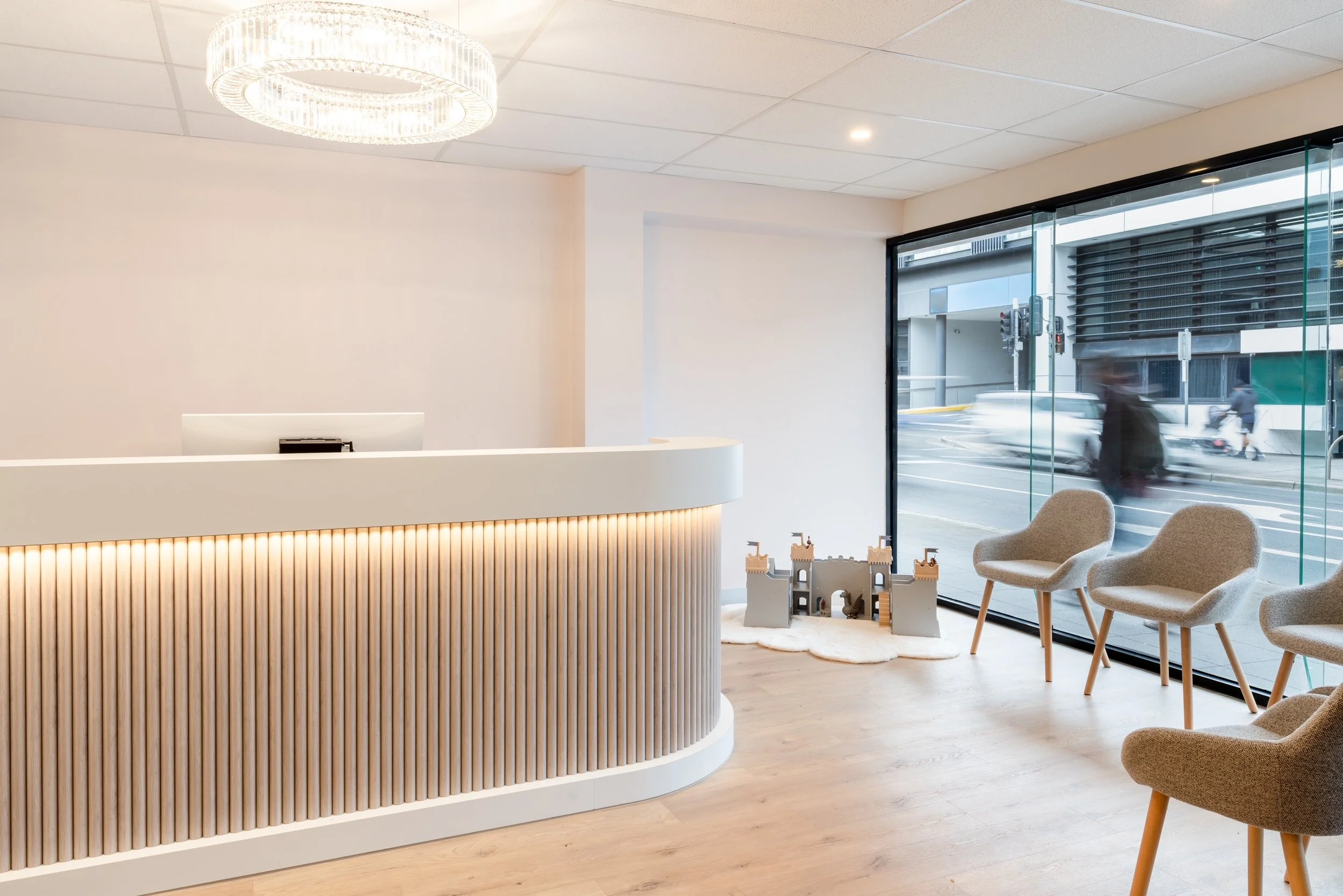 Modern reception area with a curved white desk with wooden slats, two beige chairs with wooden legs, and a large window showing city street outside with blurred cars and pedestrians.