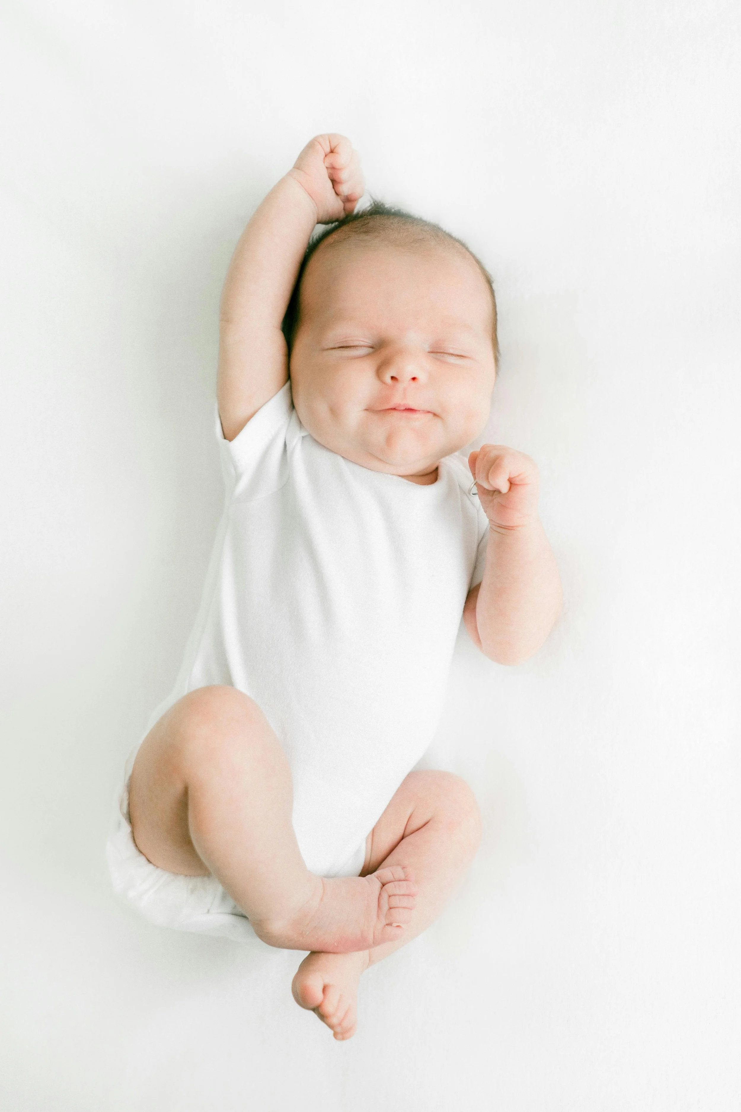 A smiling newborn baby with closed eyes, resting on a white surface, wearing a white onesie, with a hand raised near the head and minimal background.
