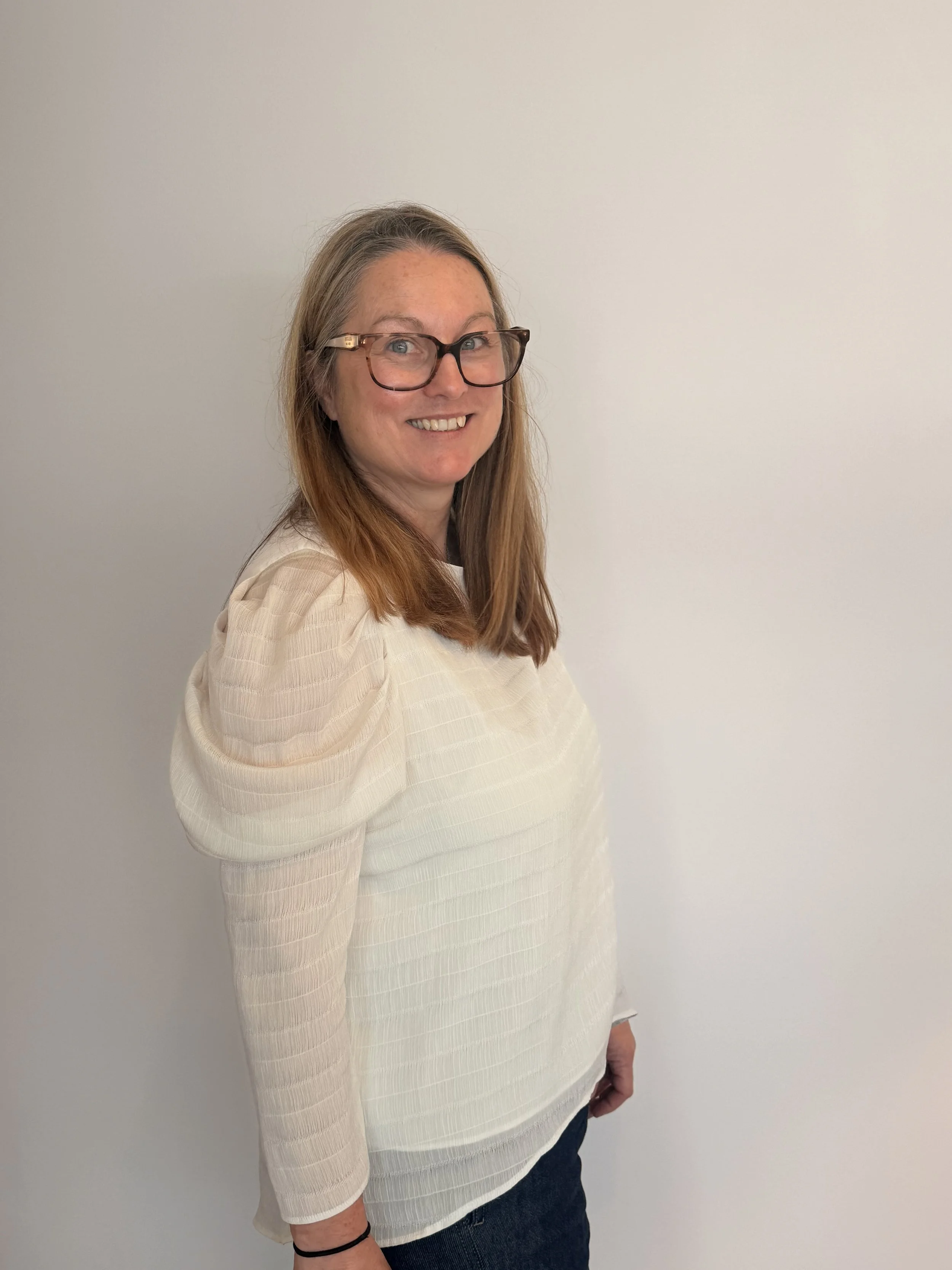Woman with long blonde hair, glasses, and a beige top standing against a plain white wall.
