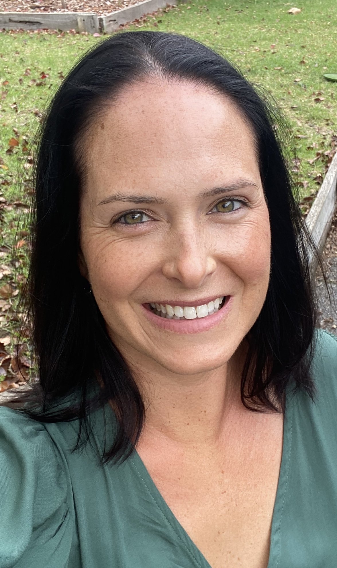 A woman with black hair, light skin with freckles, smiling outdoors with green grass and fallen leaves behind her.