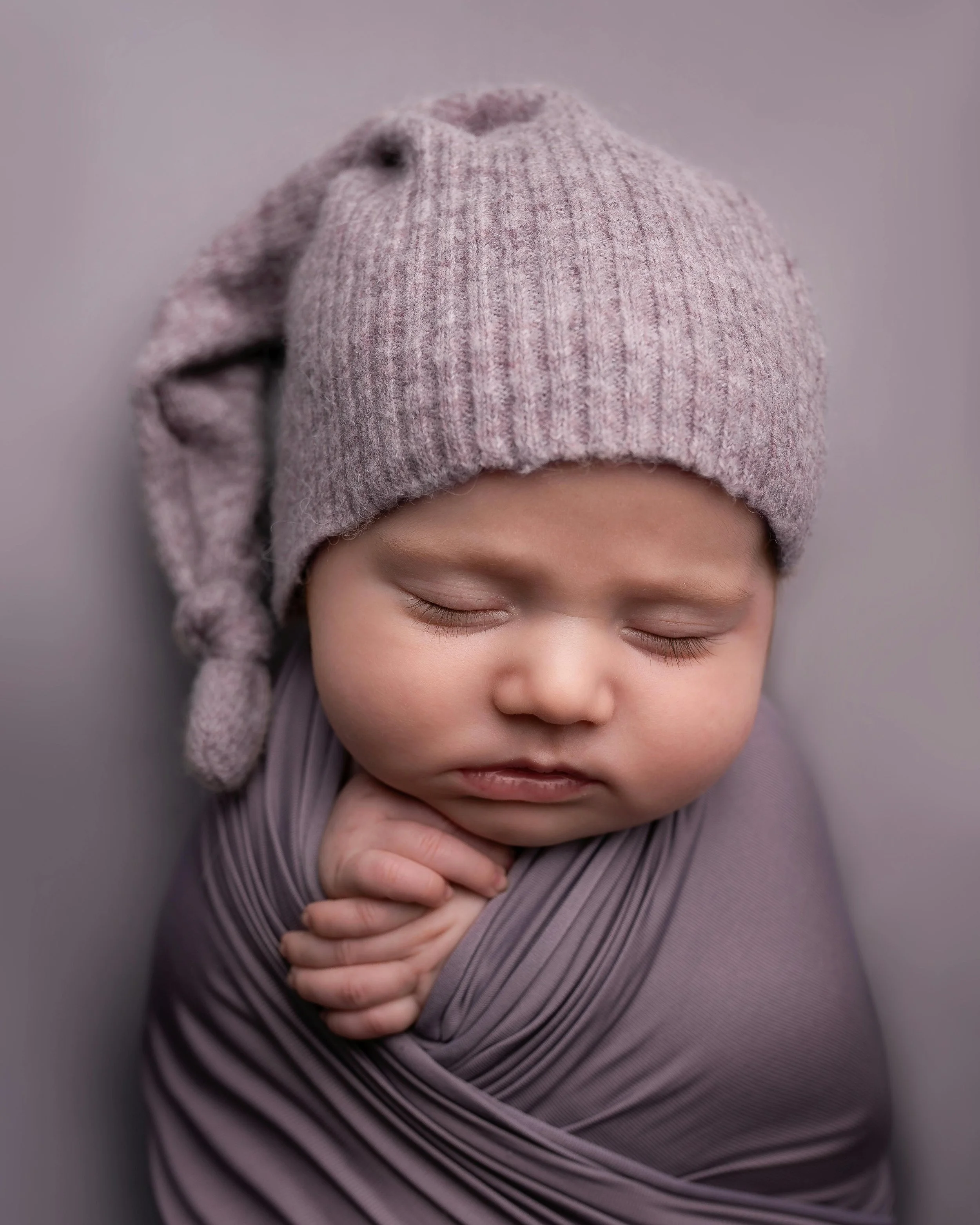 Close-up of a baby's face with eyes closed, wearing a pink knitted hat with a pom-pom on the side and a purple striped wrap.