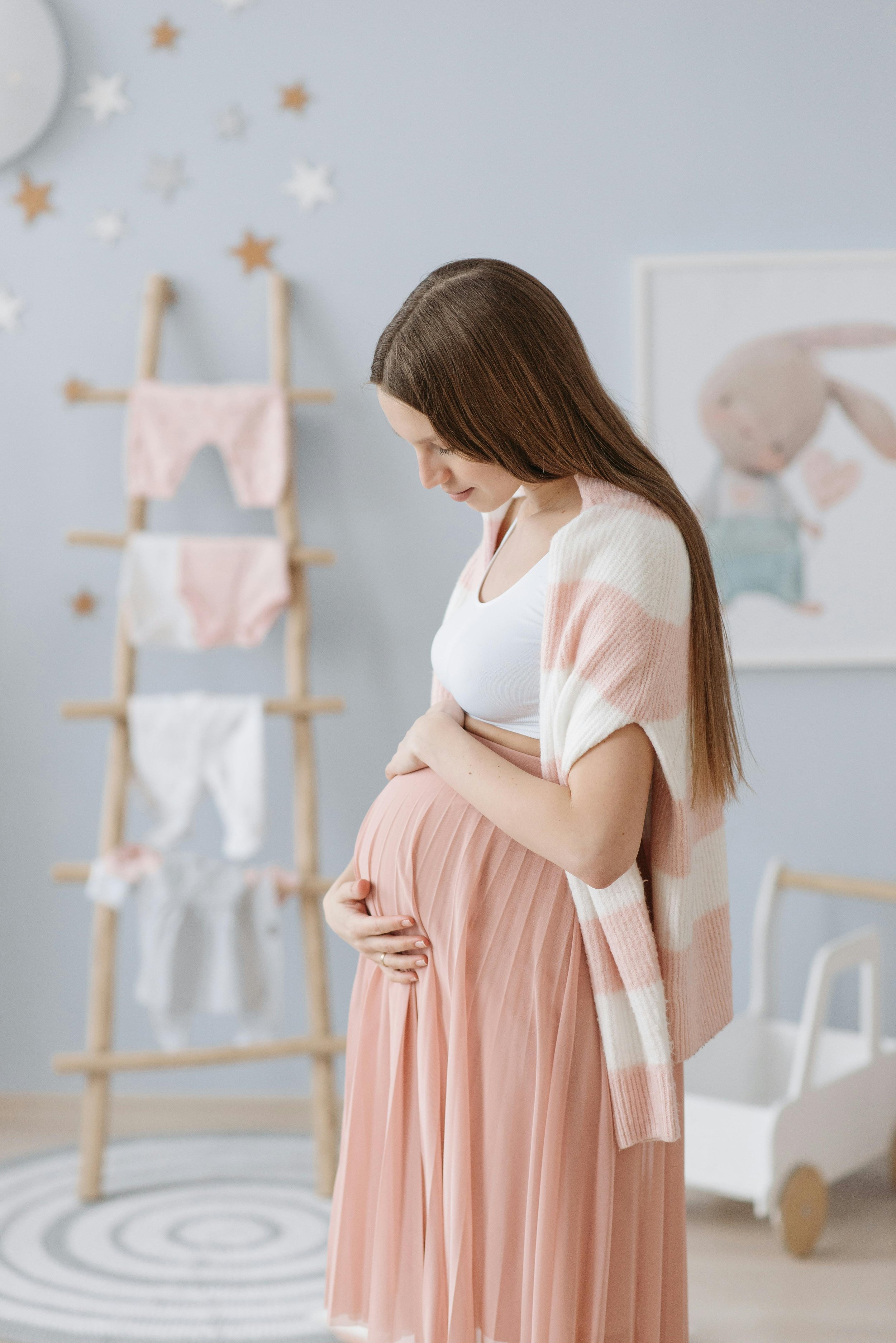 Pregnant woman in a pink skirt and white top holding her belly, standing in a nursery with star decorations and a bunny-themed wall art in the background.