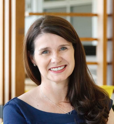 A woman with long brown hair smiling at the camera in an indoor setting.