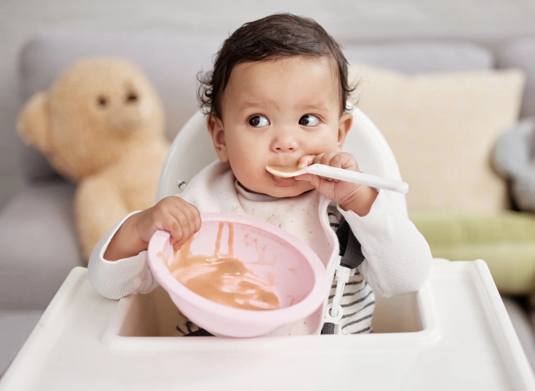 A young child sitting in a high chair with a bib, holding a spoon in their mouth while eating, with a partially eaten bowl of food in front of them and a plush toy in the background.