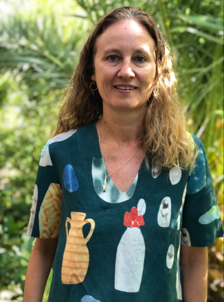 Woman with long, wavy hair smiling outdoors in a lush green environment, wearing a patterned shirt with pottery and drink illustrations.
