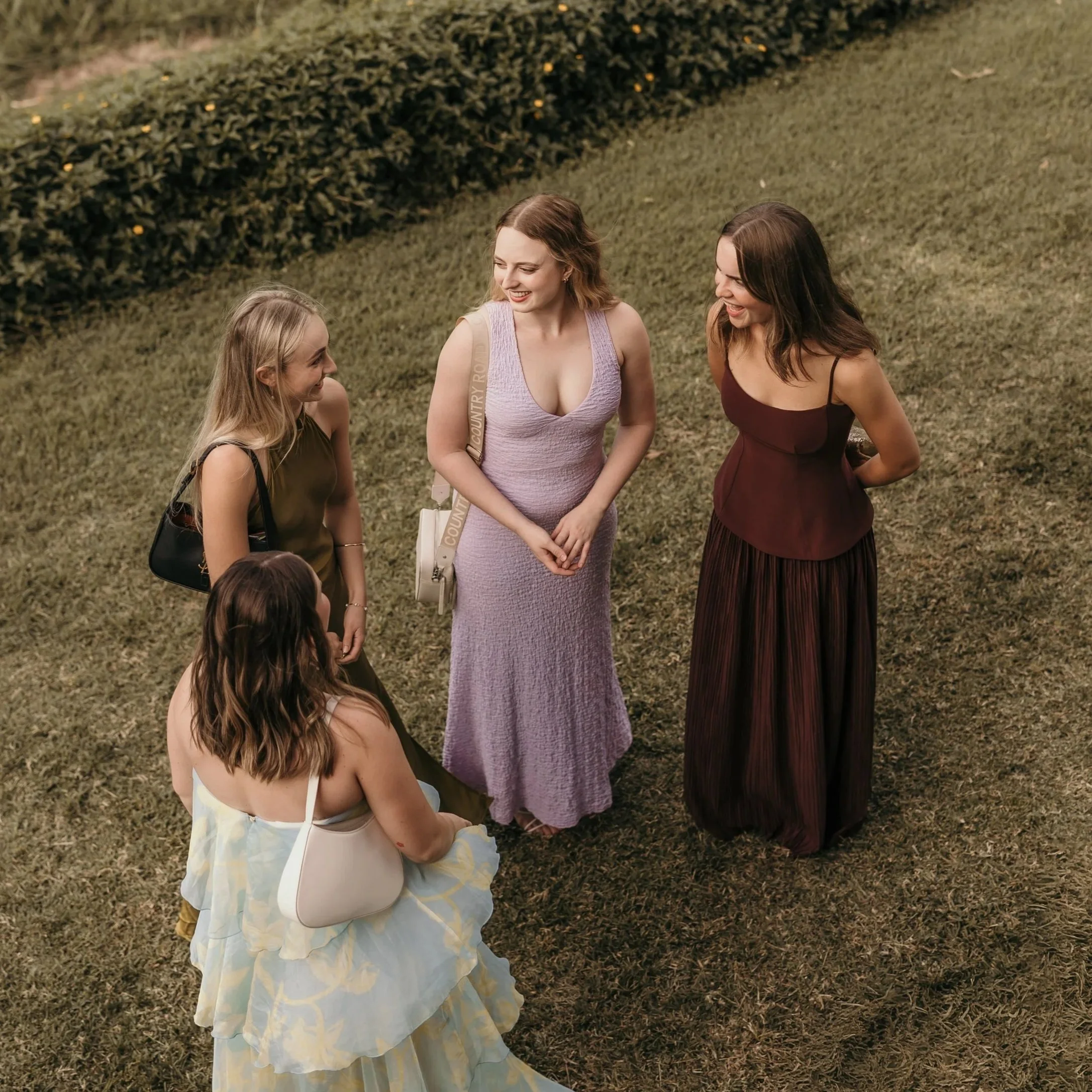 Three women and a girl are standing outside on grass, smiling and talking to each other. The woman in the middle is wearing a purple dress, the woman on the right is wearing a dark red dress, and the young girl on the left is wearing a light-colored dress with a floral pattern. They seem to be enjoying a friendly conversation.