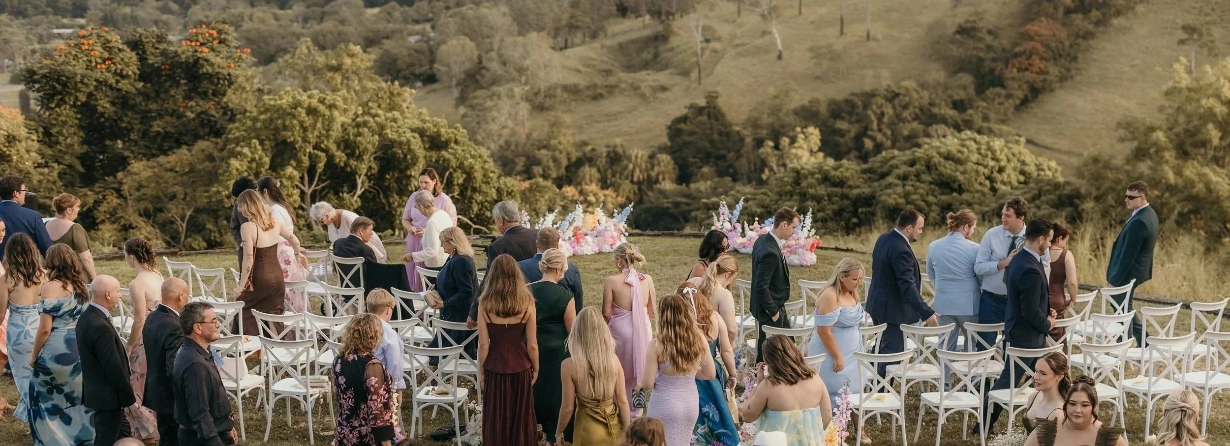 Outdoor wedding ceremony with guests seated on white chairs, floral arrangements, and a scenic green hillside in the background.