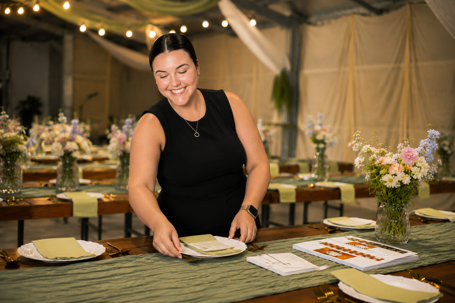 Woman in black dress setting a formal dining table with flowers and place settings in a decorated tent.