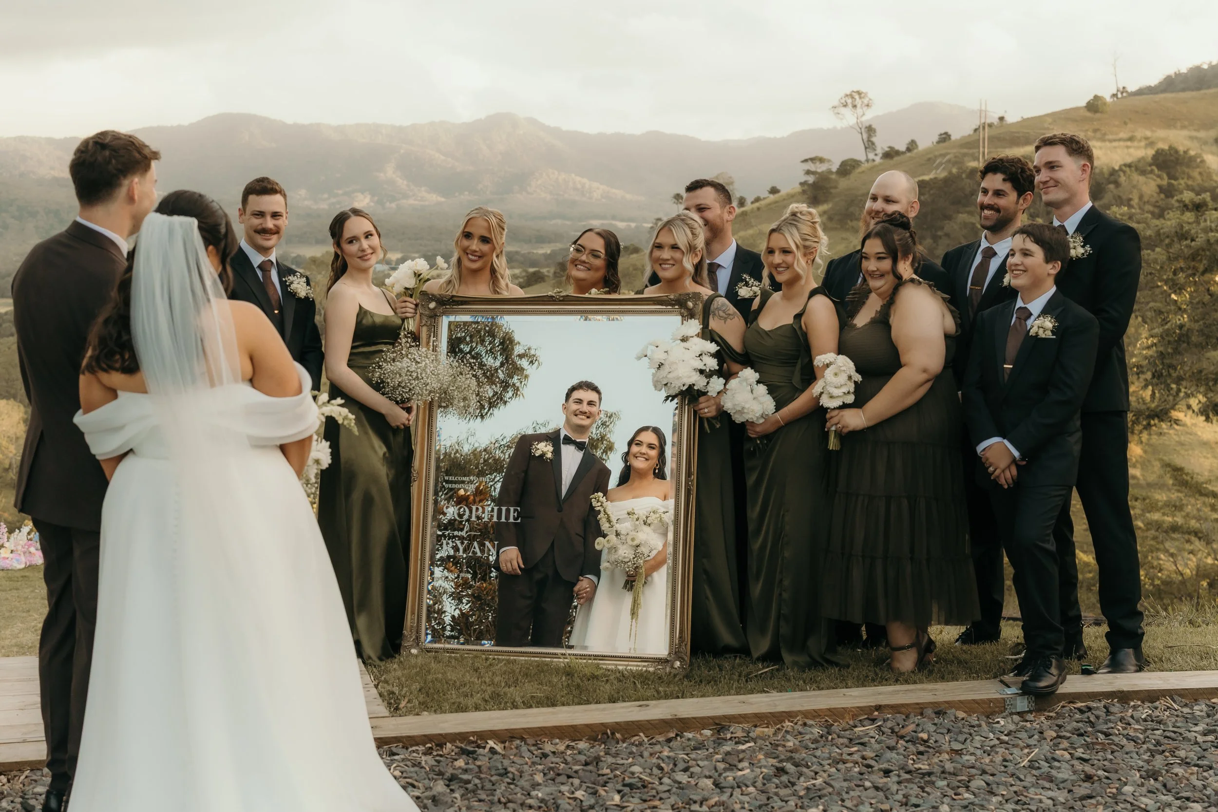 A group of wedding guests standing outdoors in a scenic mountain setting, gathered around a large framed mirror that reflects a bride and groom smiling.