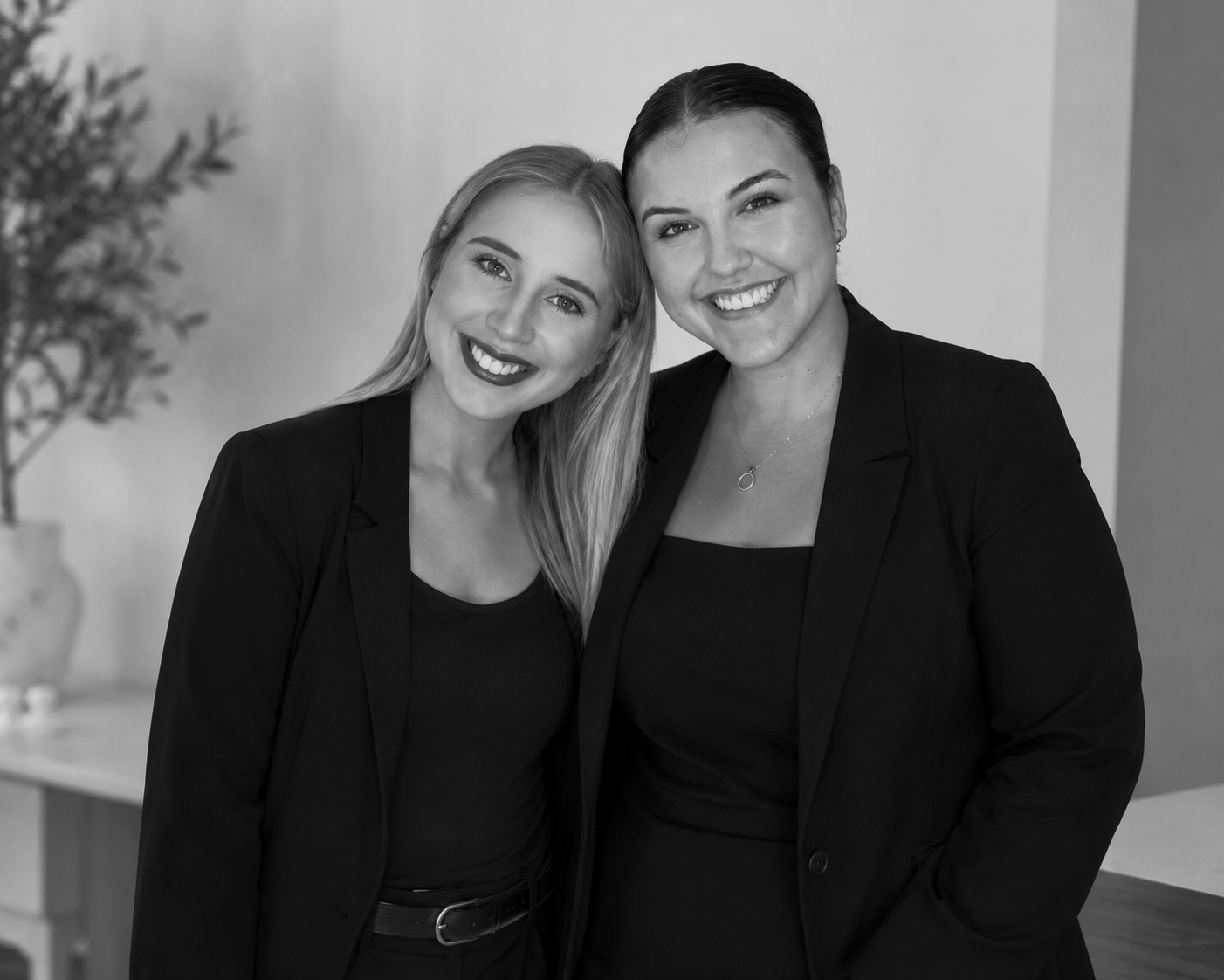 Two women with light skin smiling and standing close together, wearing black blazers and black tops in an indoor setting.