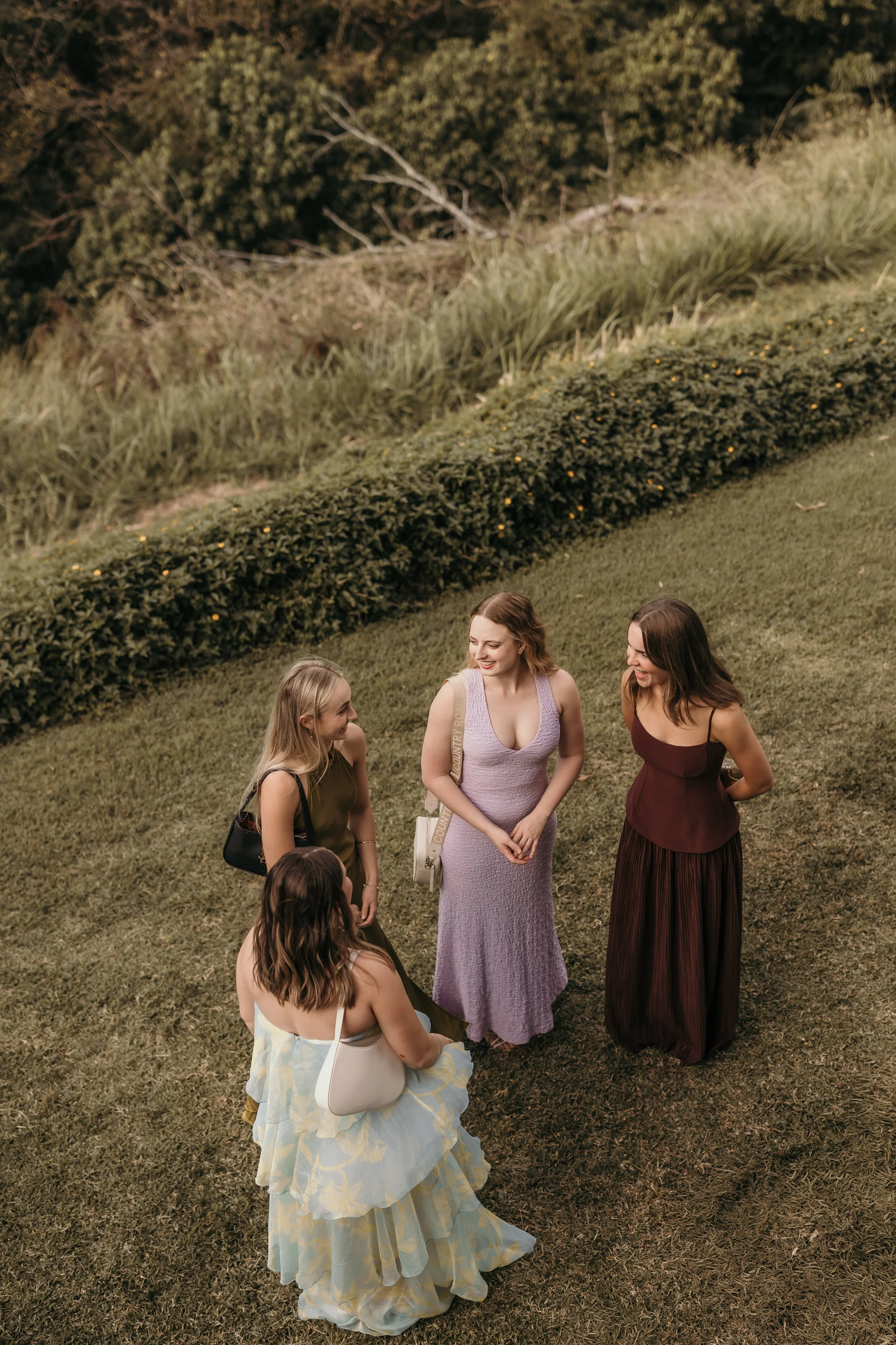 A group of women standing and sitting on grass, conversing and smiling outdoors in front of a bush and trees.