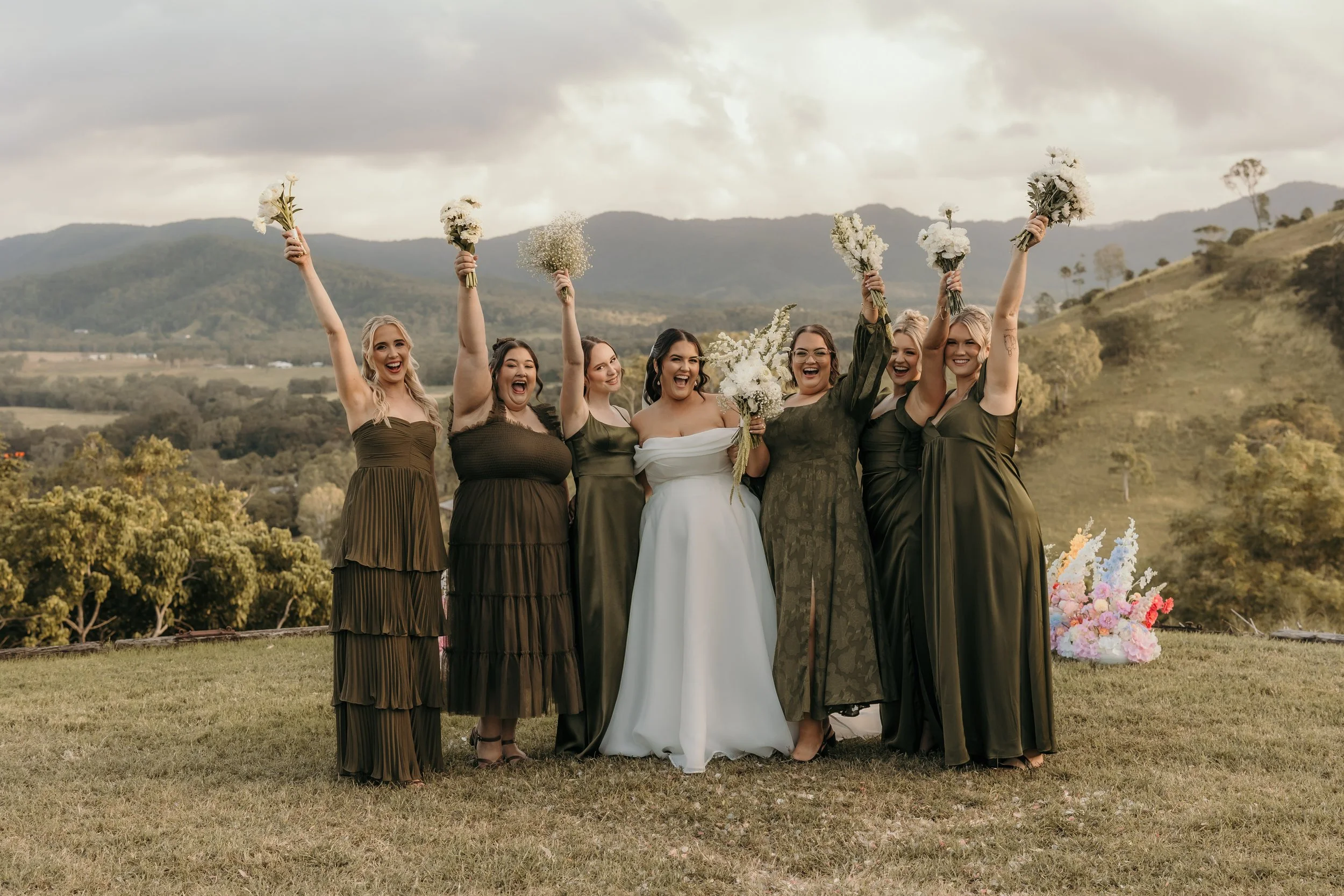A group of seven women, including a bride in a white gown, celebrating outdoors on a grassy field with rolling hills and trees in the background. They are holding bouquets of white flowers and smiling cheerfully.