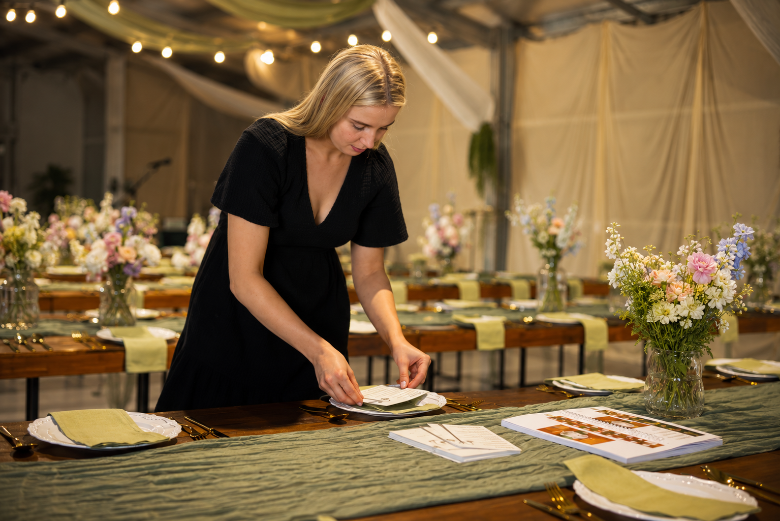 A woman in a black dress setting a table with floral centerpieces, plates, and gold utensils in a decorated event tent.