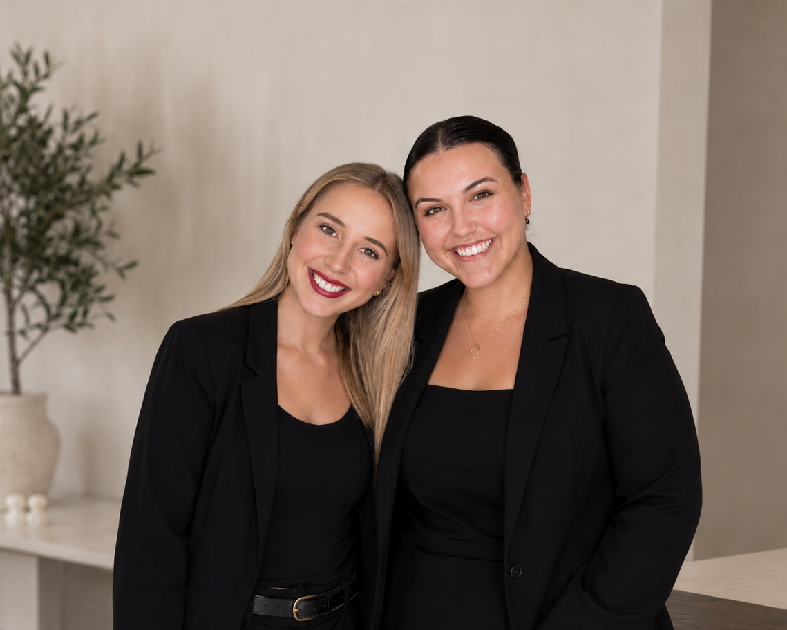 Two women in black blazers smiling and standing close together indoors with a potted plant in the background.