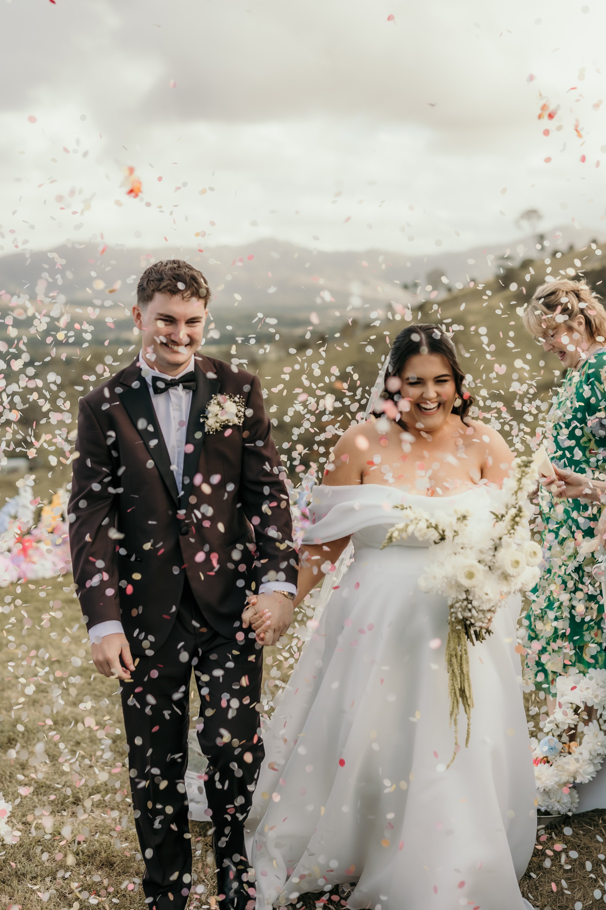 A wedding celebration outdoors with a groom and bride holding hands. The groom is in a black tuxedo with a bow tie, and the bride wears a white wedding dress holding a bouquet. They are surrounded by floating confetti and smiling, with two women standing nearby, all against a backdrop of cloudy sky and countryside.