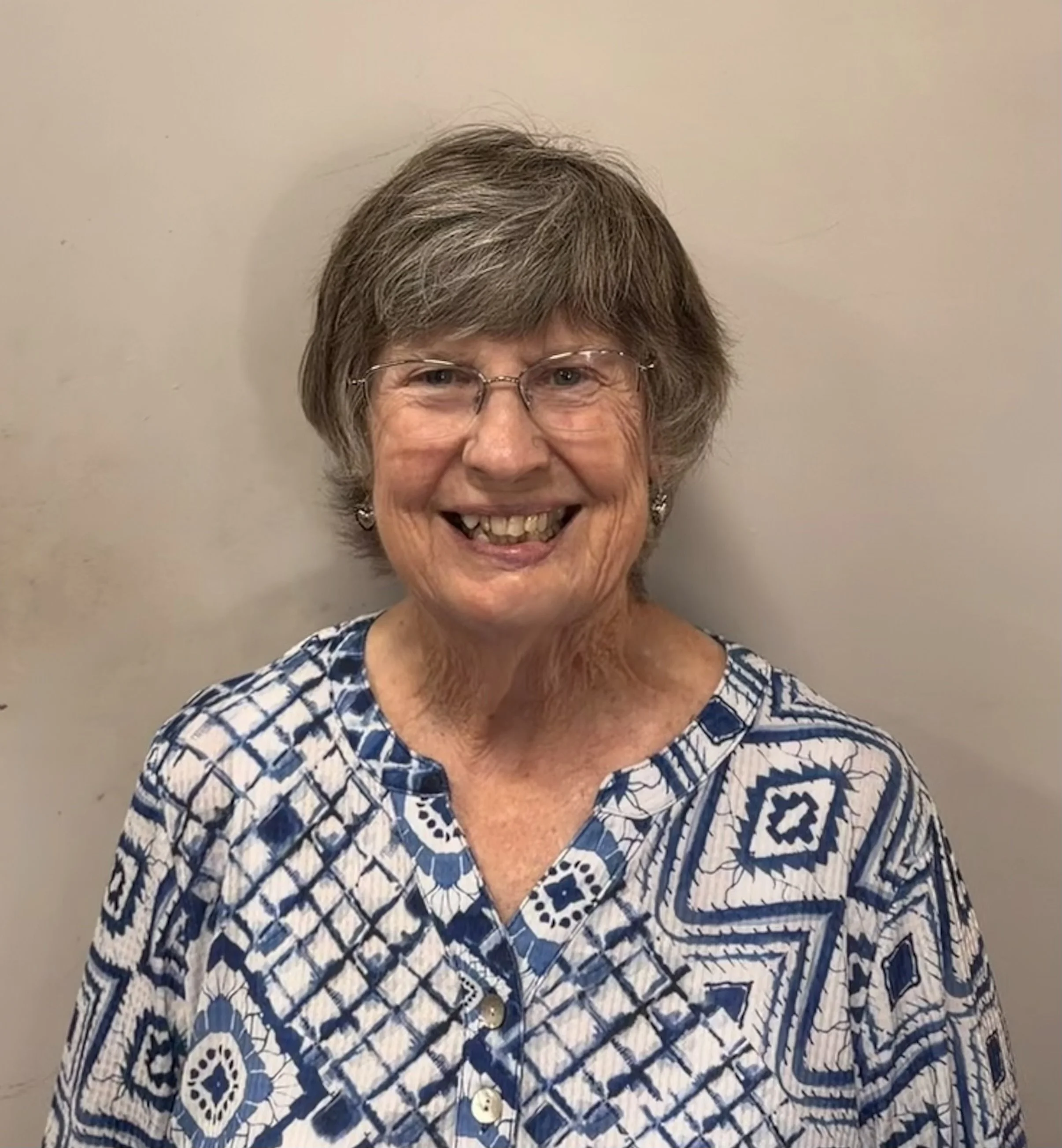 Smiling elderly woman with glasses, short gray hair, and patterned blue and white blouse standing against a plain light-colored wall.