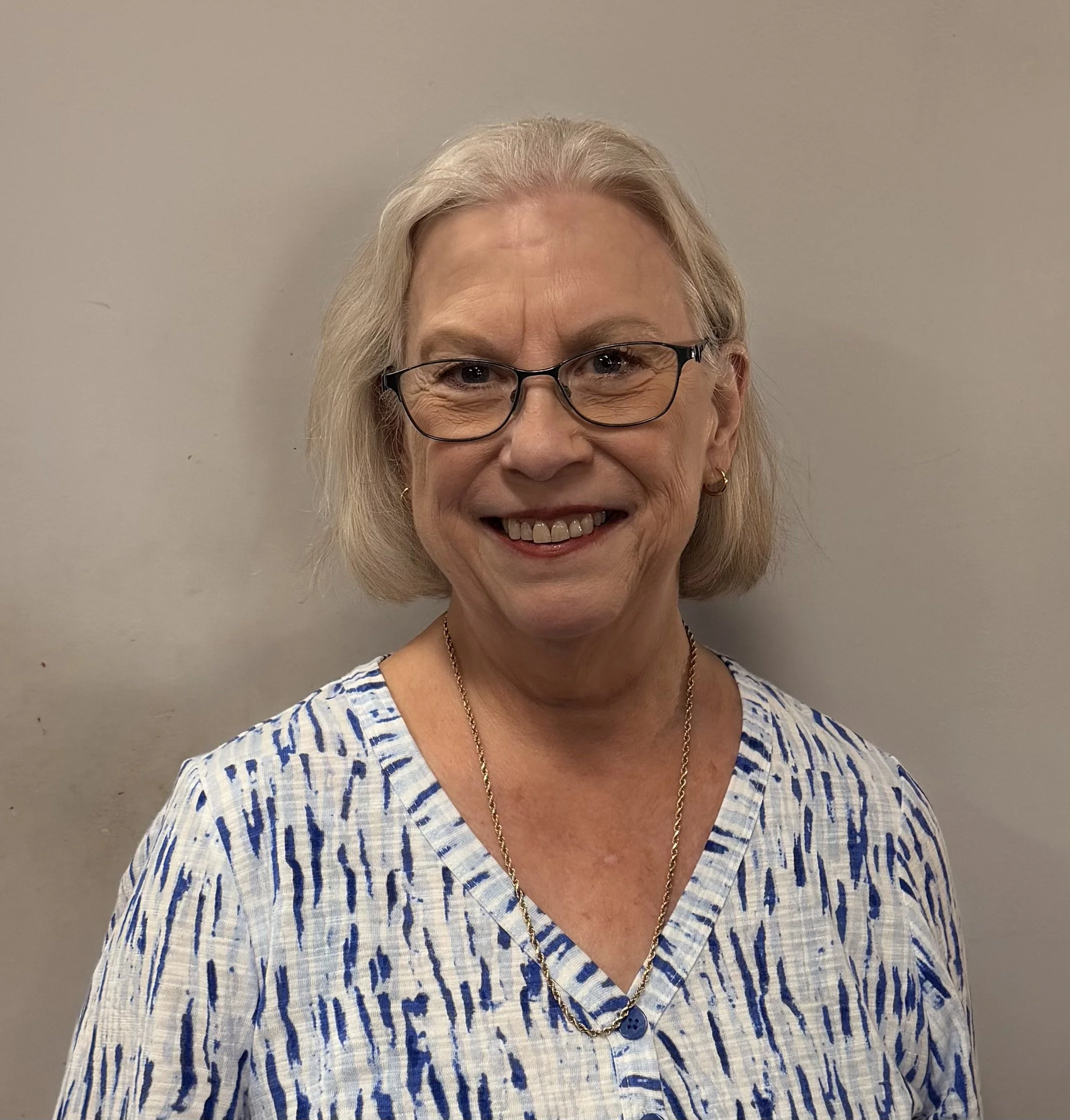 A smiling elderly woman with blonde hair, glasses, and gold jewelry, wearing a patterned blouse, standing against a plain wall.