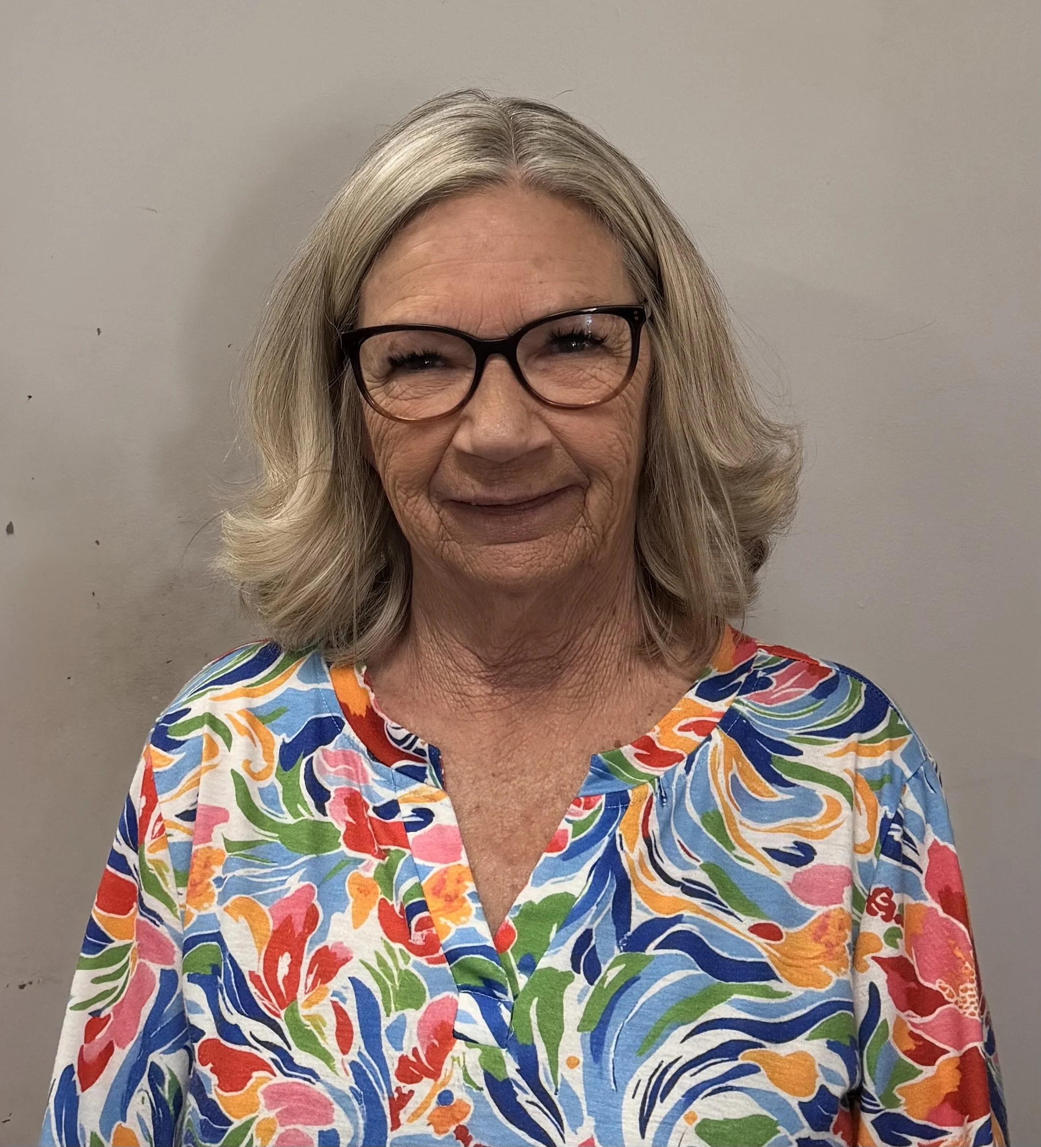 A smiling middle-aged woman with blonde hair, glasses, and a colorful, patterned blouse standing against a plain, light-colored wall.